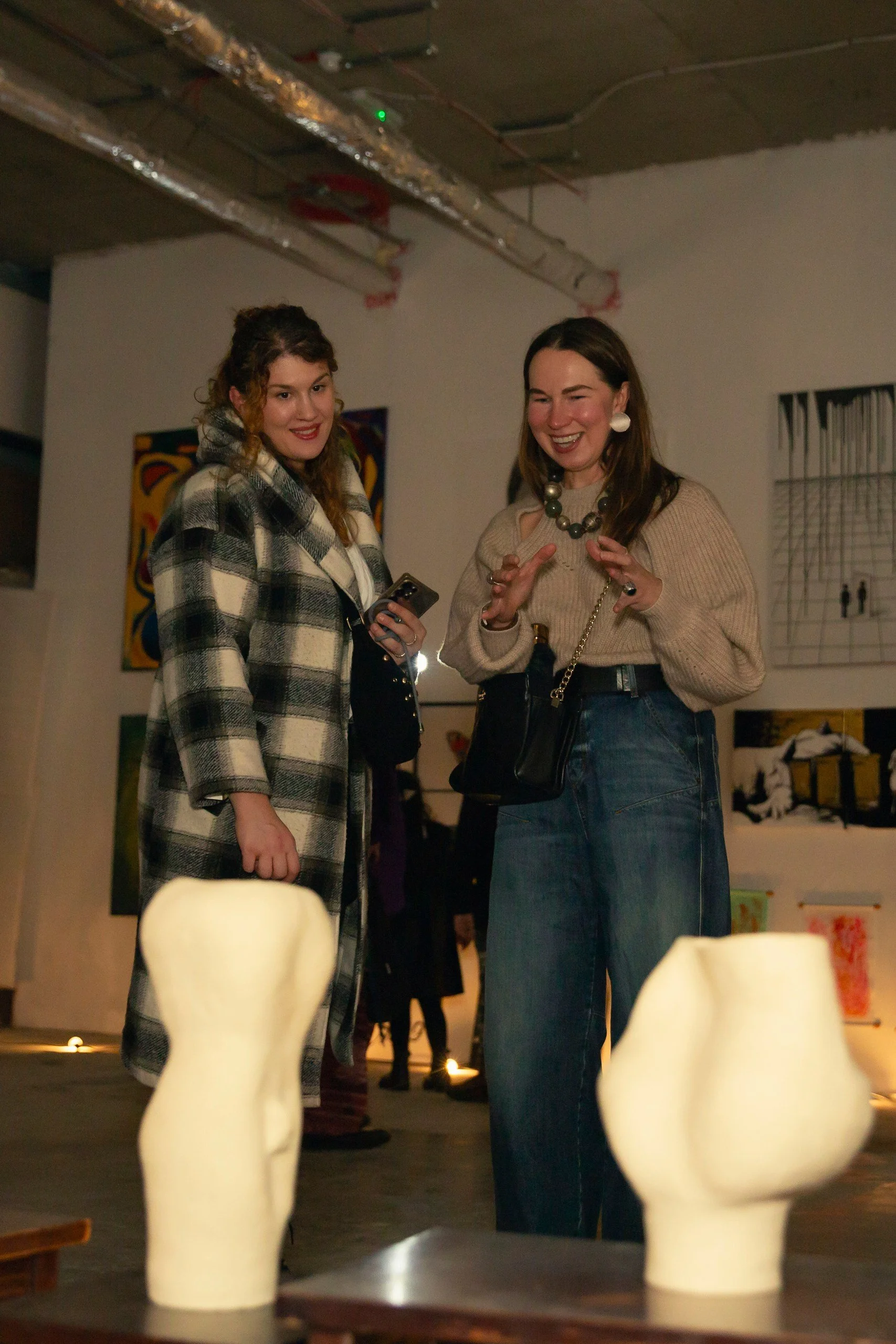 LuSee talking to an interested client during Duality show an Art.Fait art gallery, with abstract paintings on the wall behind them and white sculptural handmade vases on a table in the foreground.