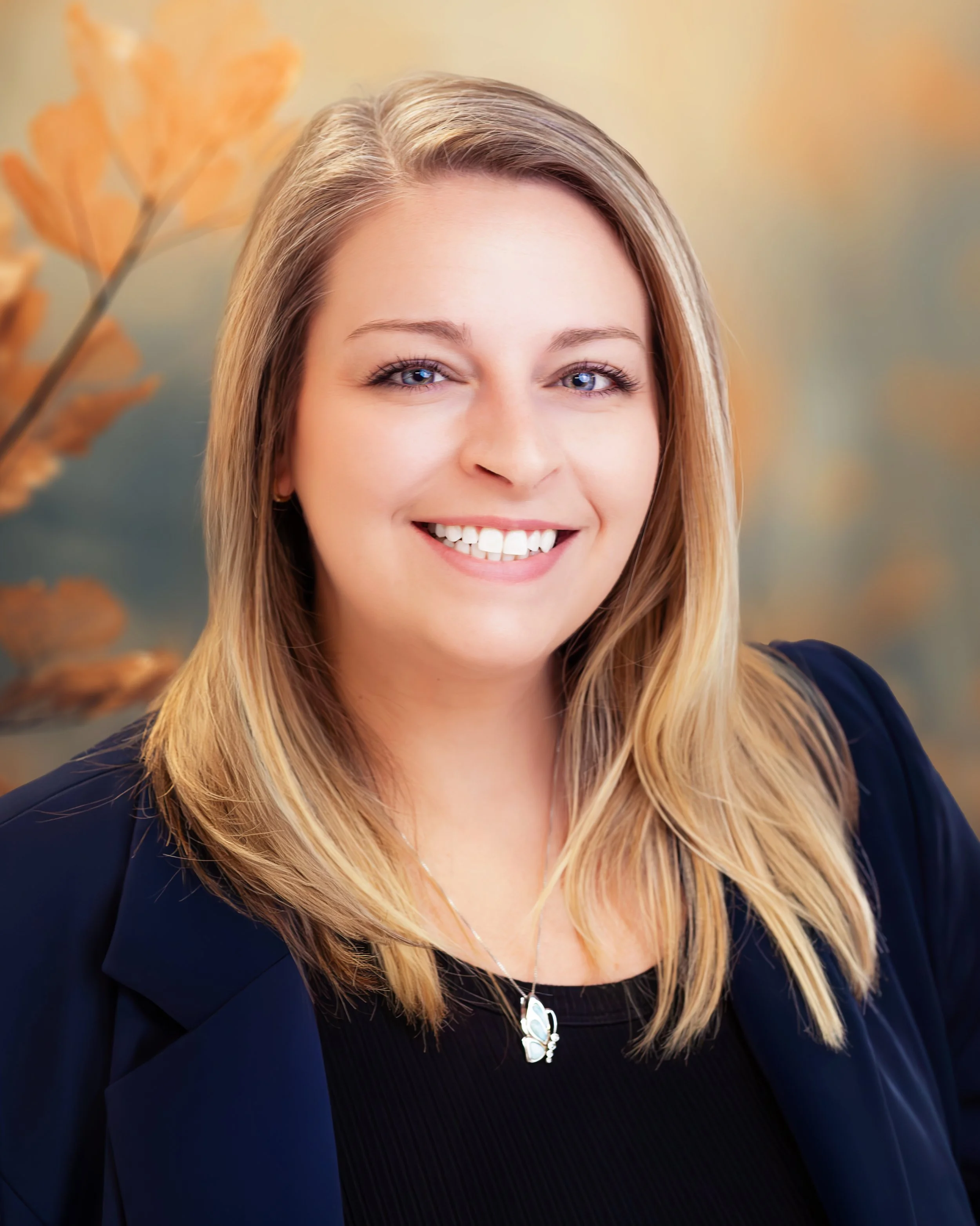 A smiling woman with blonde hair, wearing a black top and a dark blazer, with a butterfly necklace, against a blurred autumn background.