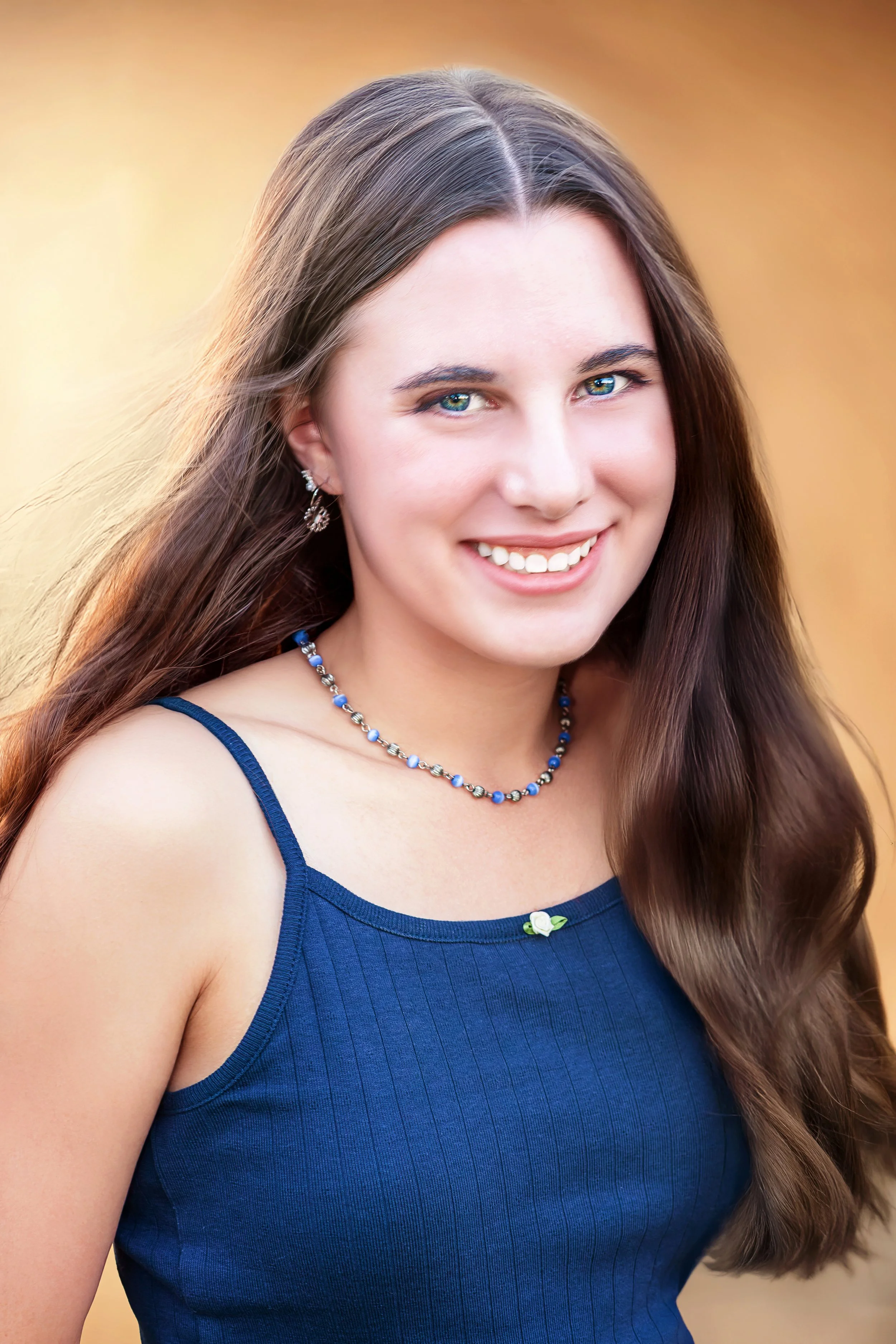 A young woman with long brown hair, blue eyes, and a bright smile wearing a blue sleeveless top, a necklace, and earrings, outdoors during daytime. Headshot Photography in Loveland, Colorado. Jeanette Fellows Photography www.fellows.photos