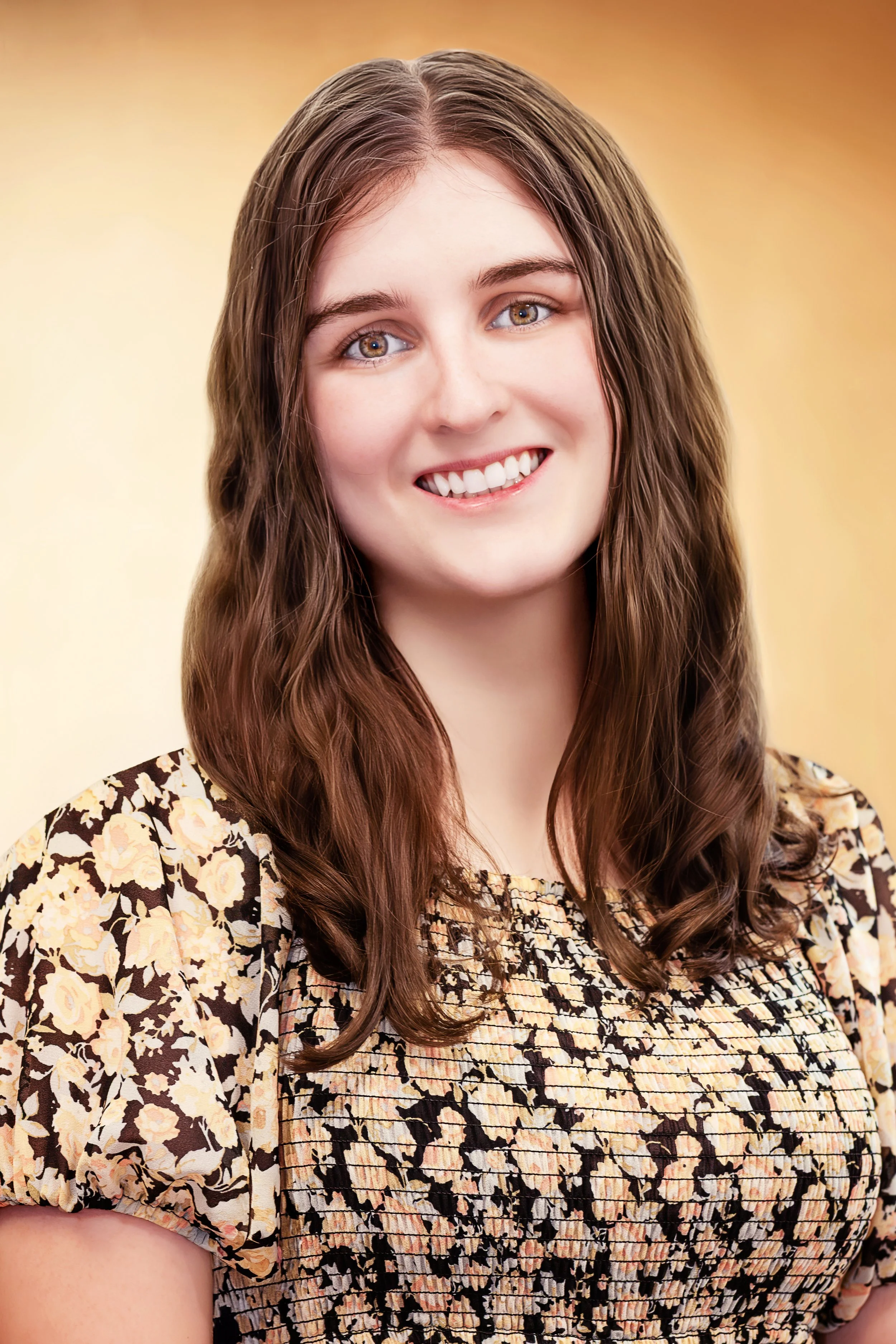 A young woman with long brown hair, smiling, wearing a patterned blouse with floral and abstract design, and a yellowish background. Headshot Photography in Loveland, Colorado. Jeanette Fellows Photography www.fellows.photos
