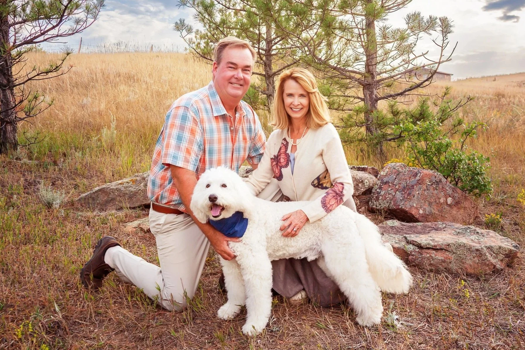 A happy couple with a large white fluffy dog in an outdoor grassy area with trees and hills in the background during daytime. Jeanette Fellows Photography www.fellows.photos
