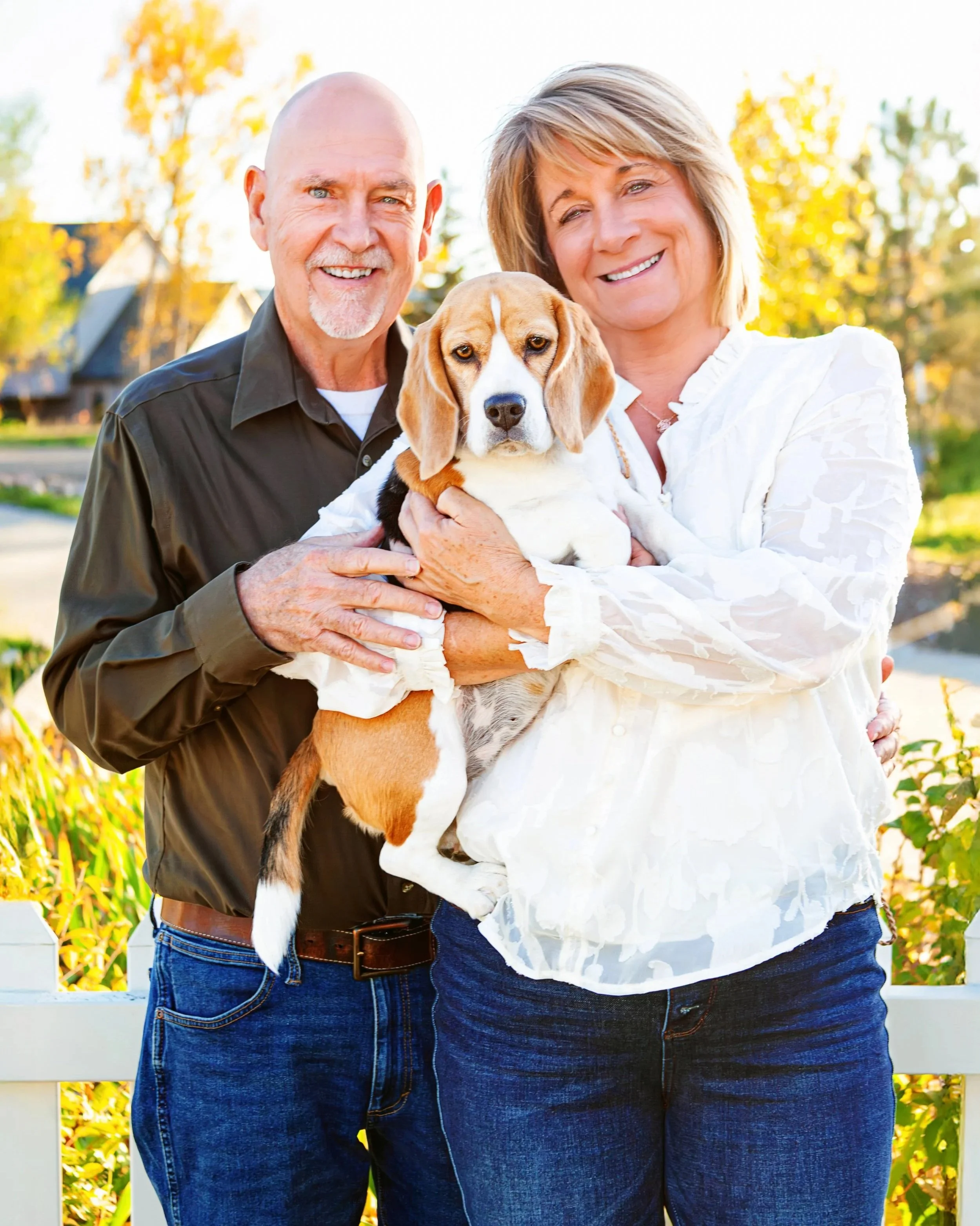 A smiling man and woman holding a beagle puppy outdoors during fall, with trees and a house in the background. Jeanette Fellows Photography www.fellows.photos