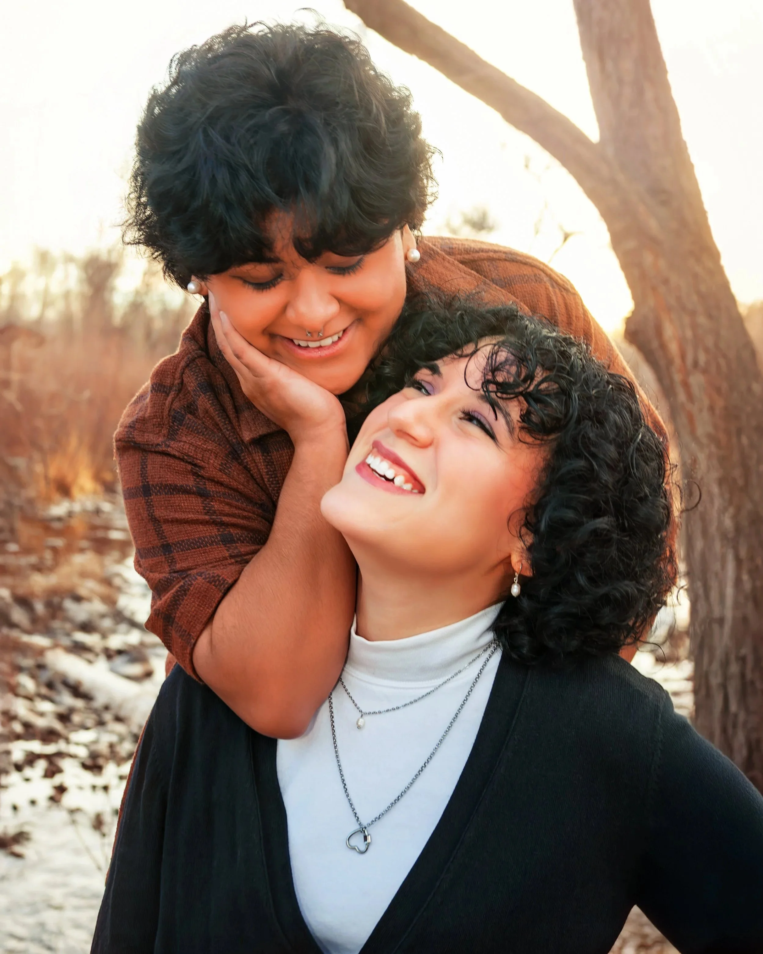 Two women, one with short dark hair and the other with curly hair, are outdoors during sunset. They are smiling and embracing, with the woman with short hair gently holding the face of the other. Trees and a hint of snow are visible in the background