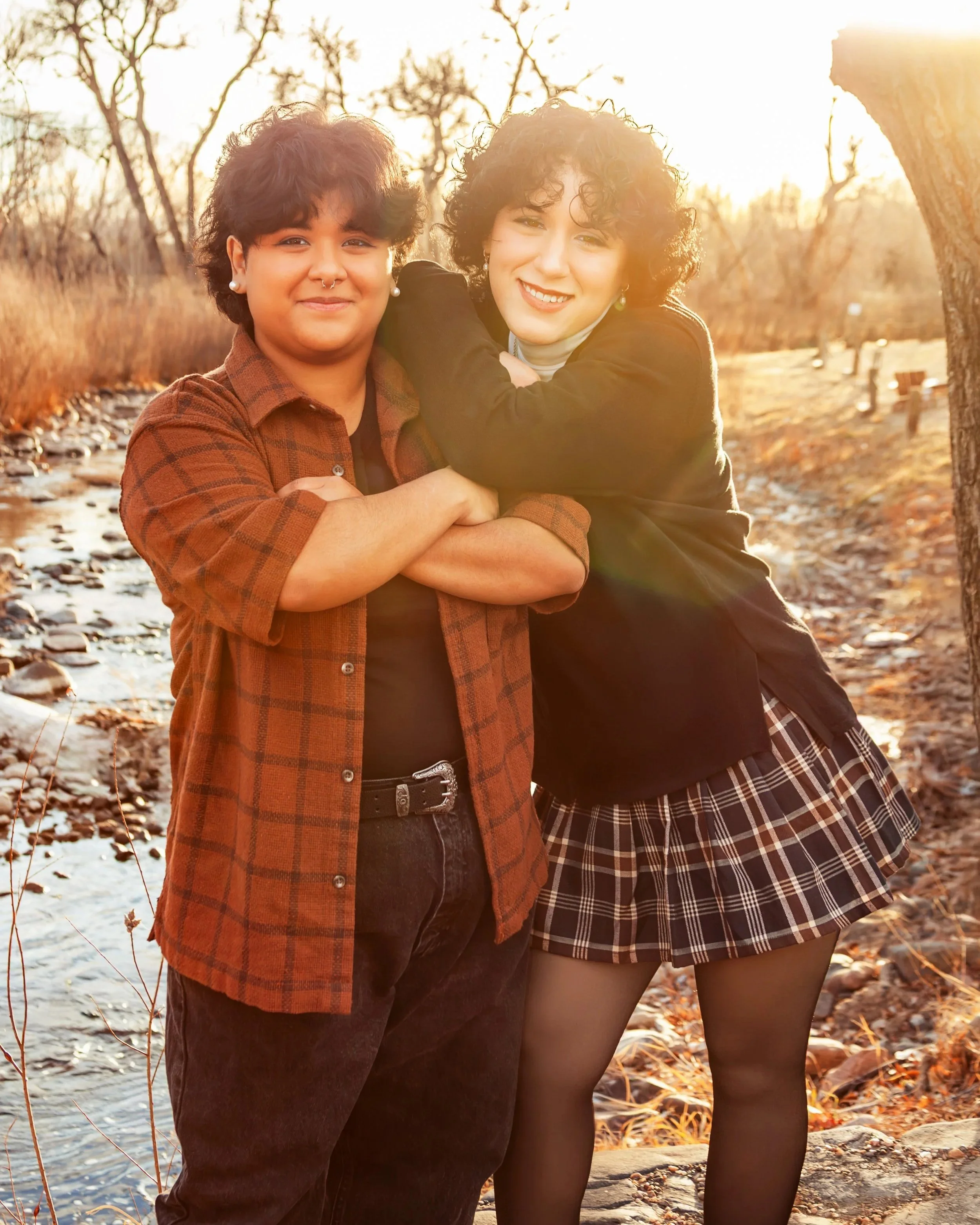 Two women standing outdoors in a creekside setting during sunset, smiling, with one embracing the other. Jeanette Fellows Photography www.fellows.photos