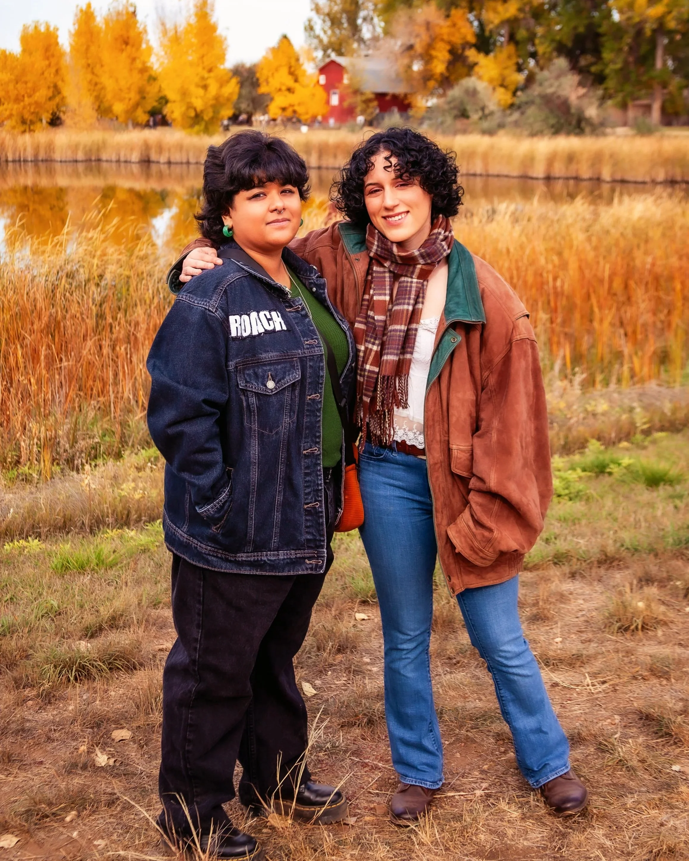 Two women standing together outdoors in fall scenery, near a lake with trees with yellow and orange leaves in the background. Jeanette Fellows Photography in Loveland, Colorado