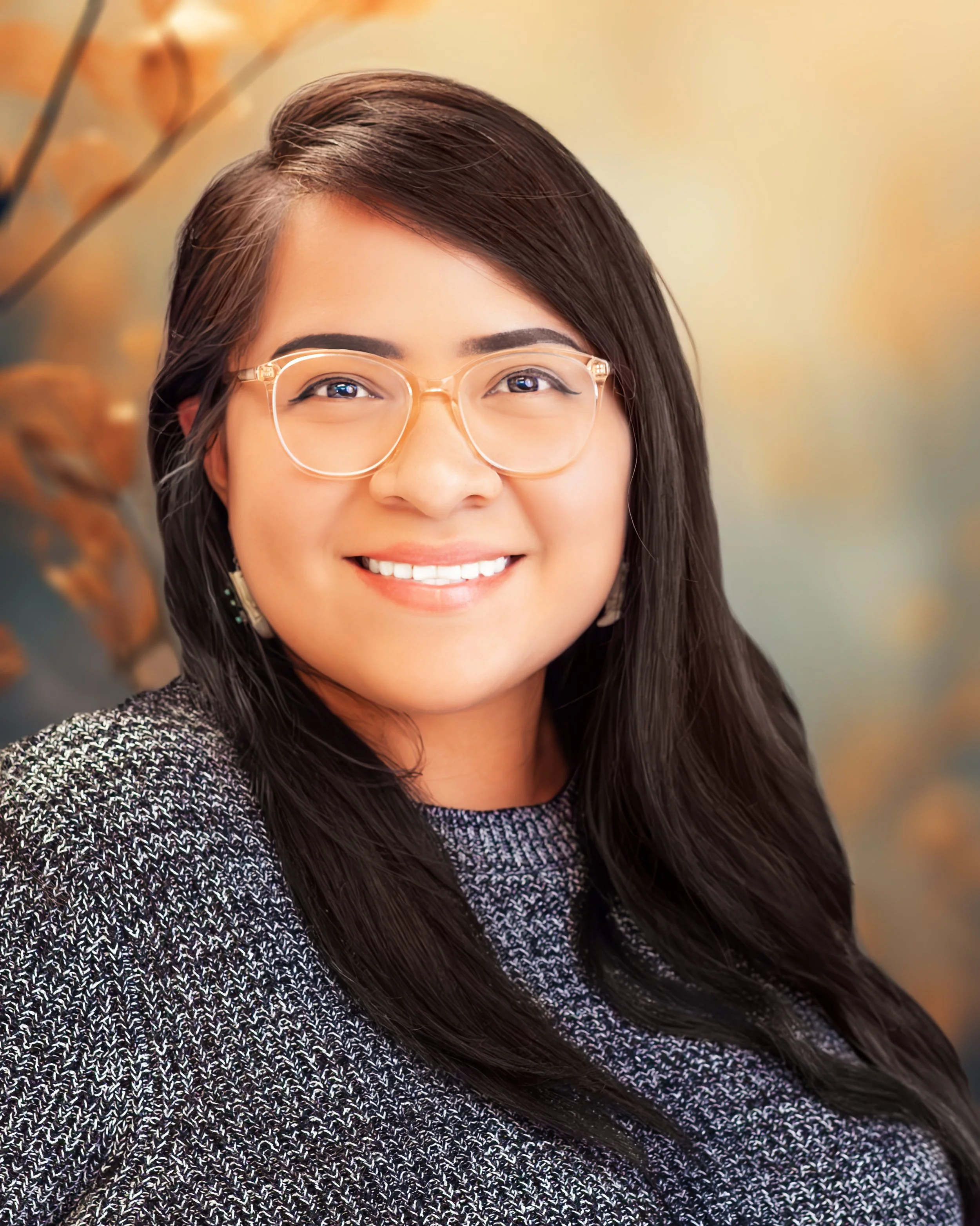 A woman with long dark hair, wearing glasses and a textured sweater, smiling indoors with a blurred autumnal background.