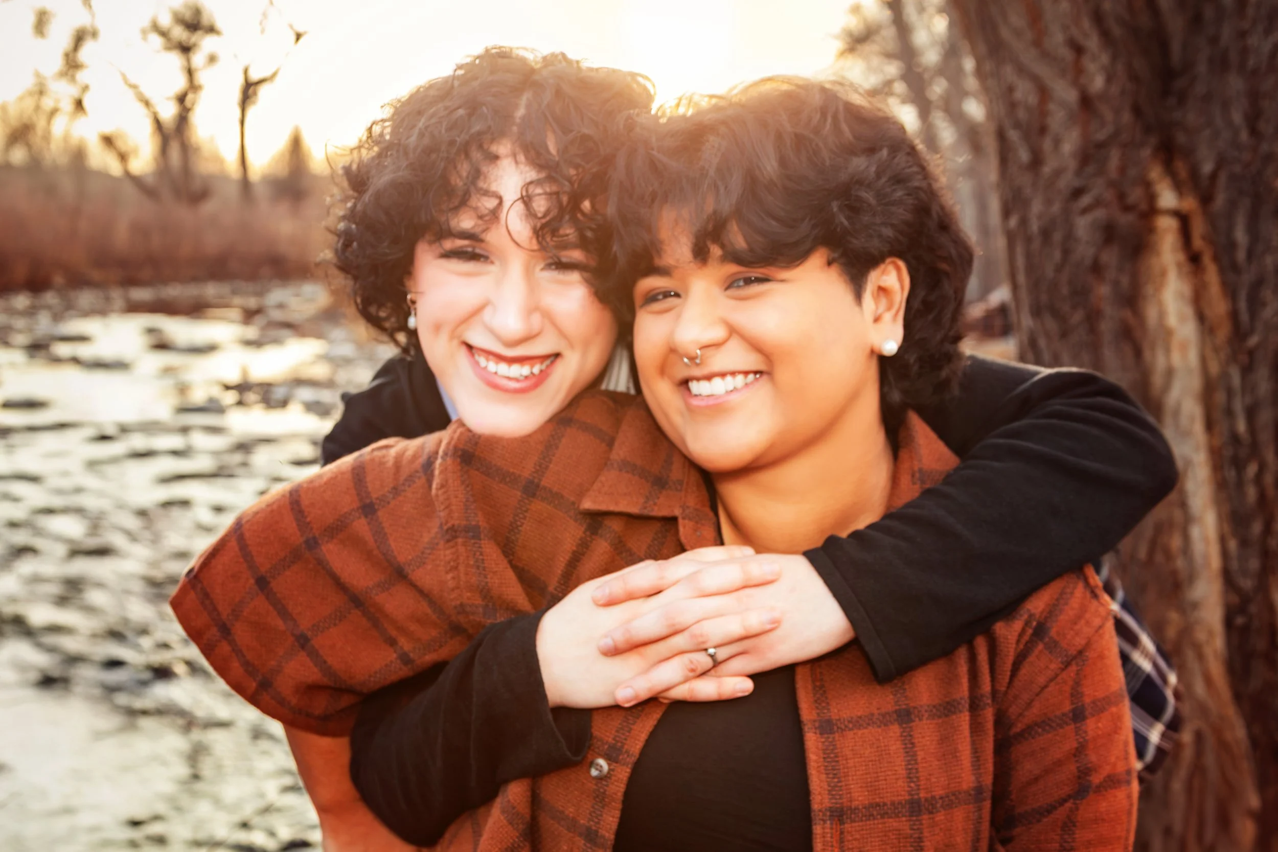 Two smiling women embracing outdoors near a river with trees in the background during sunset. Jeanette Fellows Photography www.fellows.photos