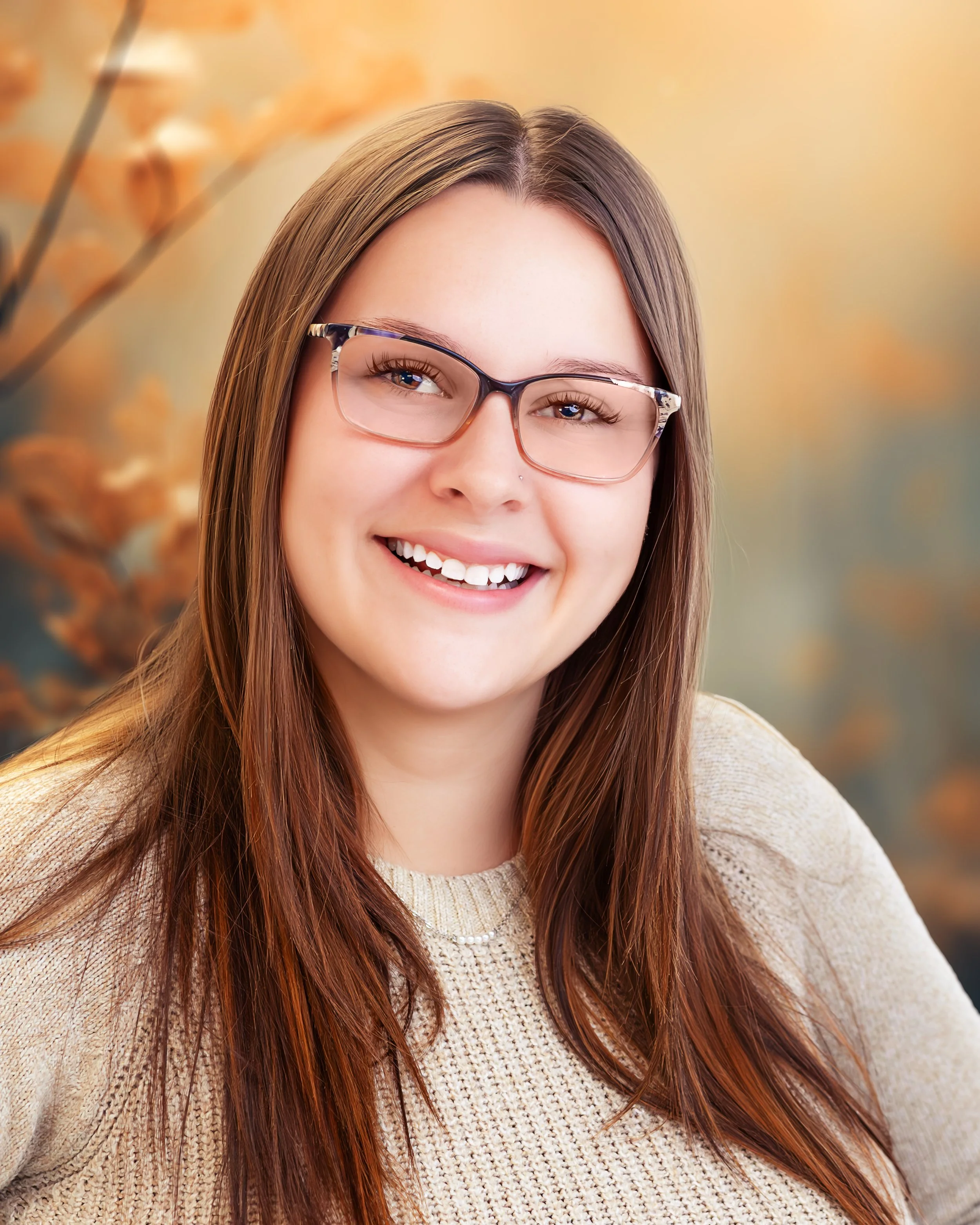 A smiling woman with long brown hair, glasses, and a beige sweater, against a blurred autumn background.