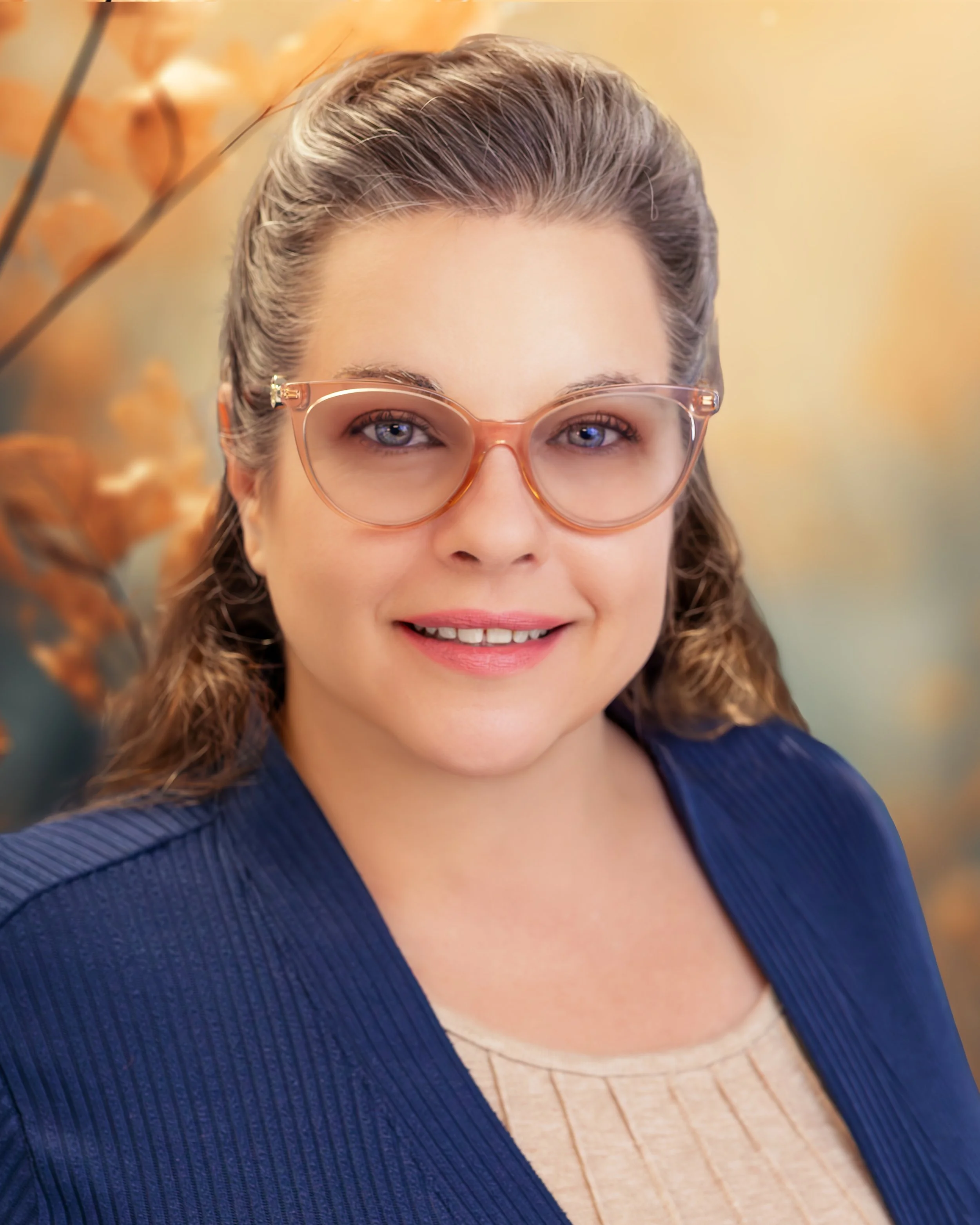 A woman with light brown, wavy hair, wearing glasses with a light pink frame, a navy blue blazer, and a beige top, smiling at the camera with a blurred floral background.
