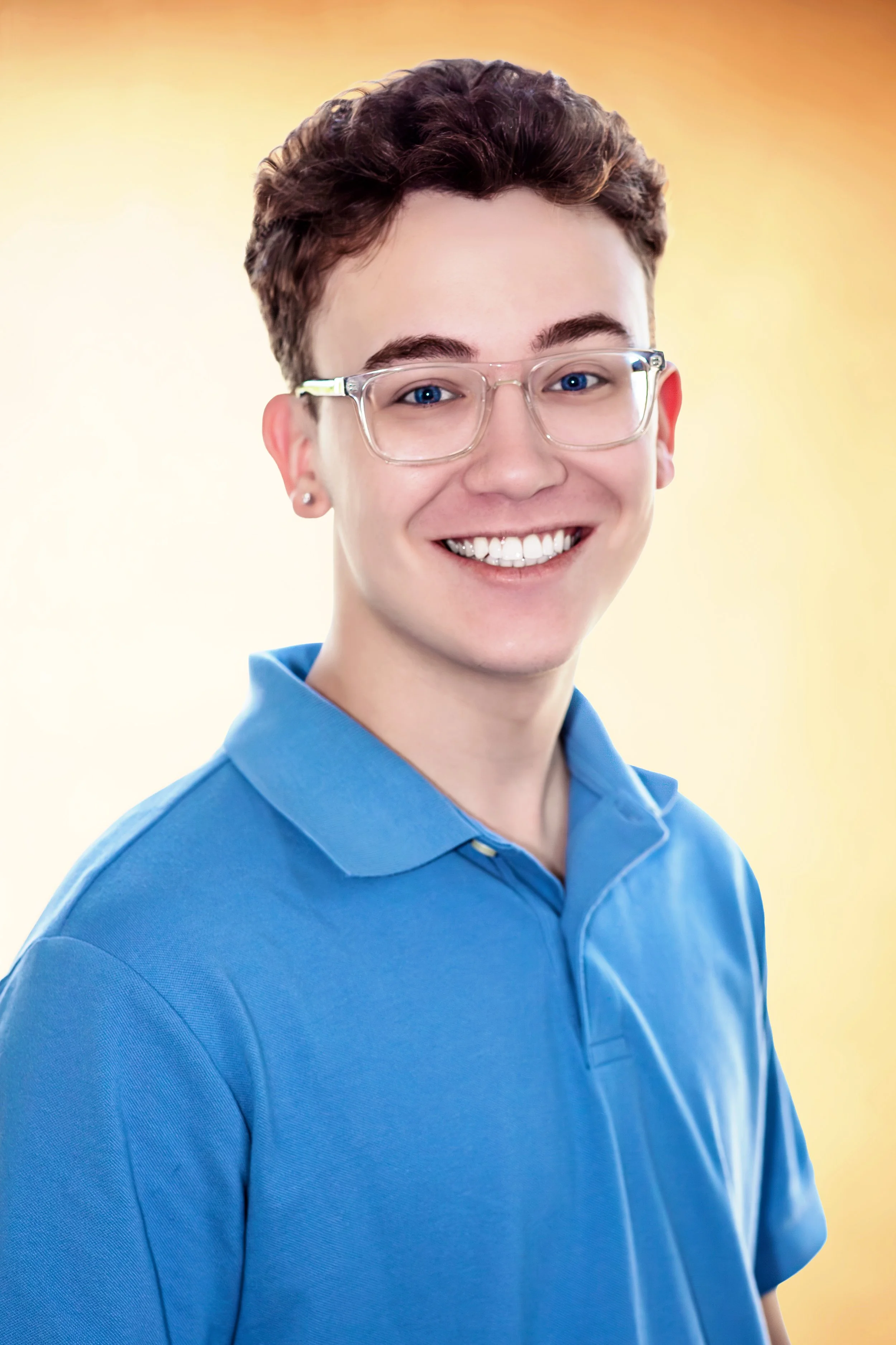 A young person with short curly hair, glasses, and earrings, smiling and wearing a blue polo shirt against a warm yellow background. Headshot Photography in Loveland, Colorado. Jeanette Fellows Photography www.fellows.photos