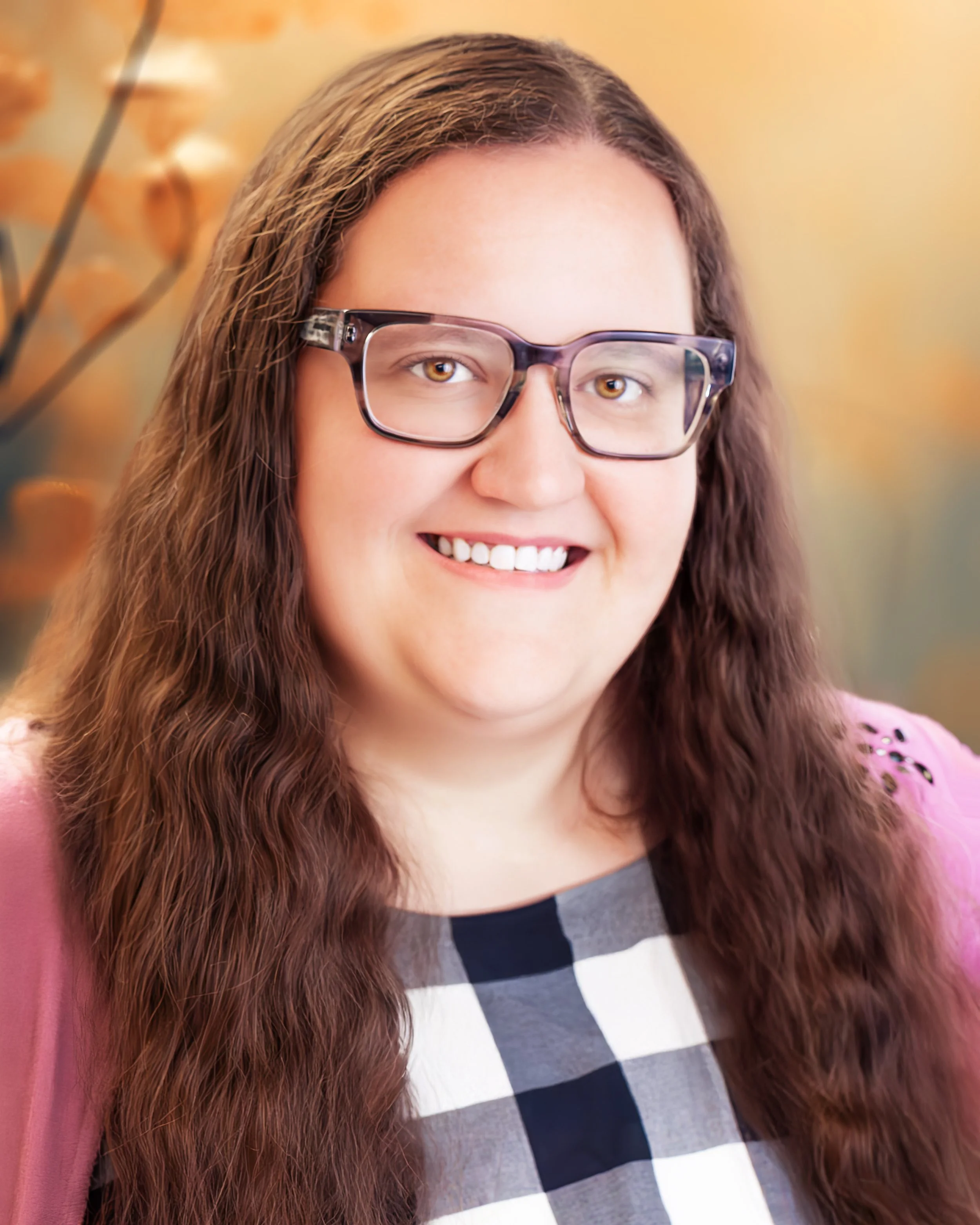 Close-up of woman with long wavy brown hair, glasses, and a patterned top, smiling against a blurred background.