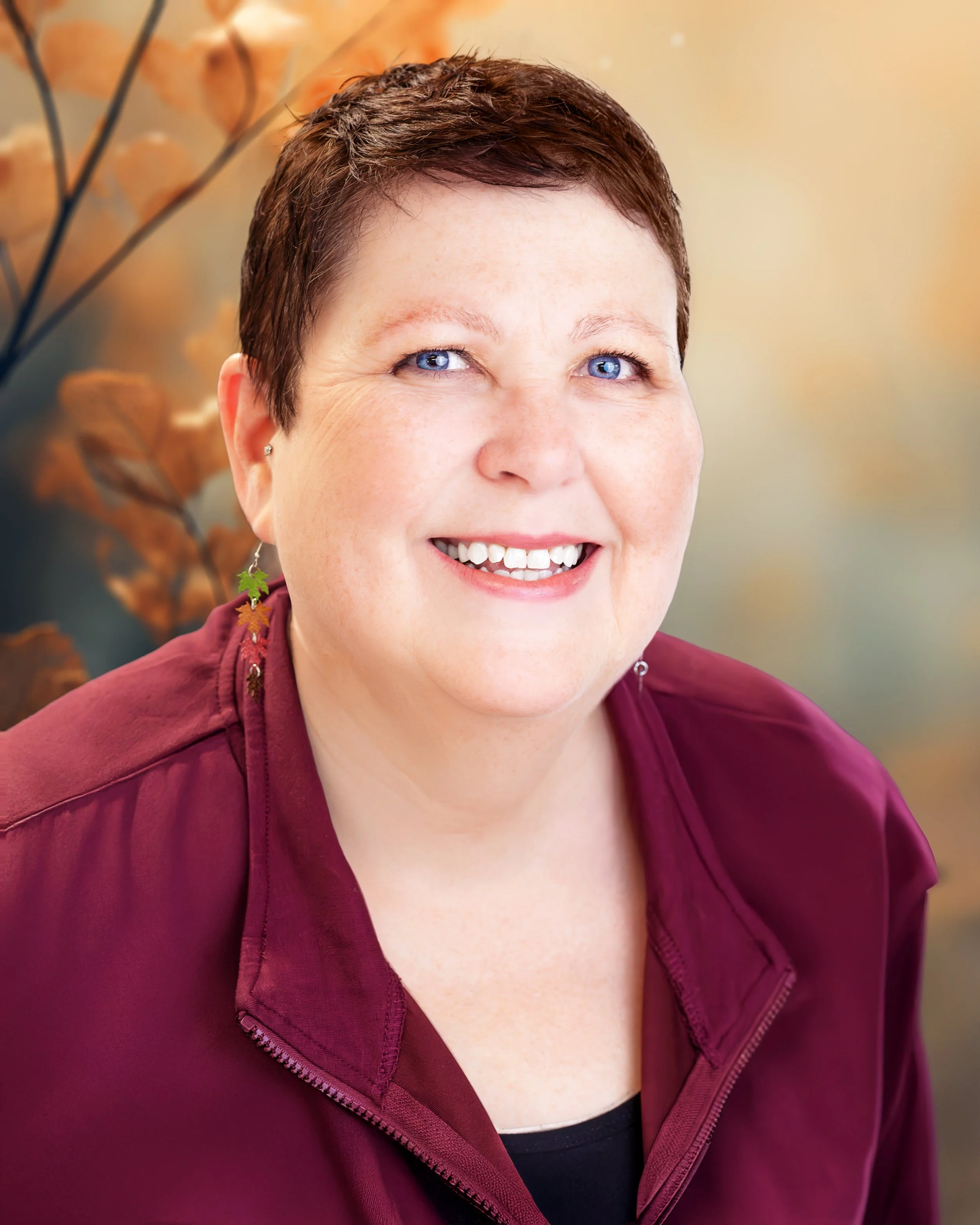 A smiling woman with short brown hair and blue eyes, wearing earrings and a burgundy zip-up jacket, against a blurred autumn background.