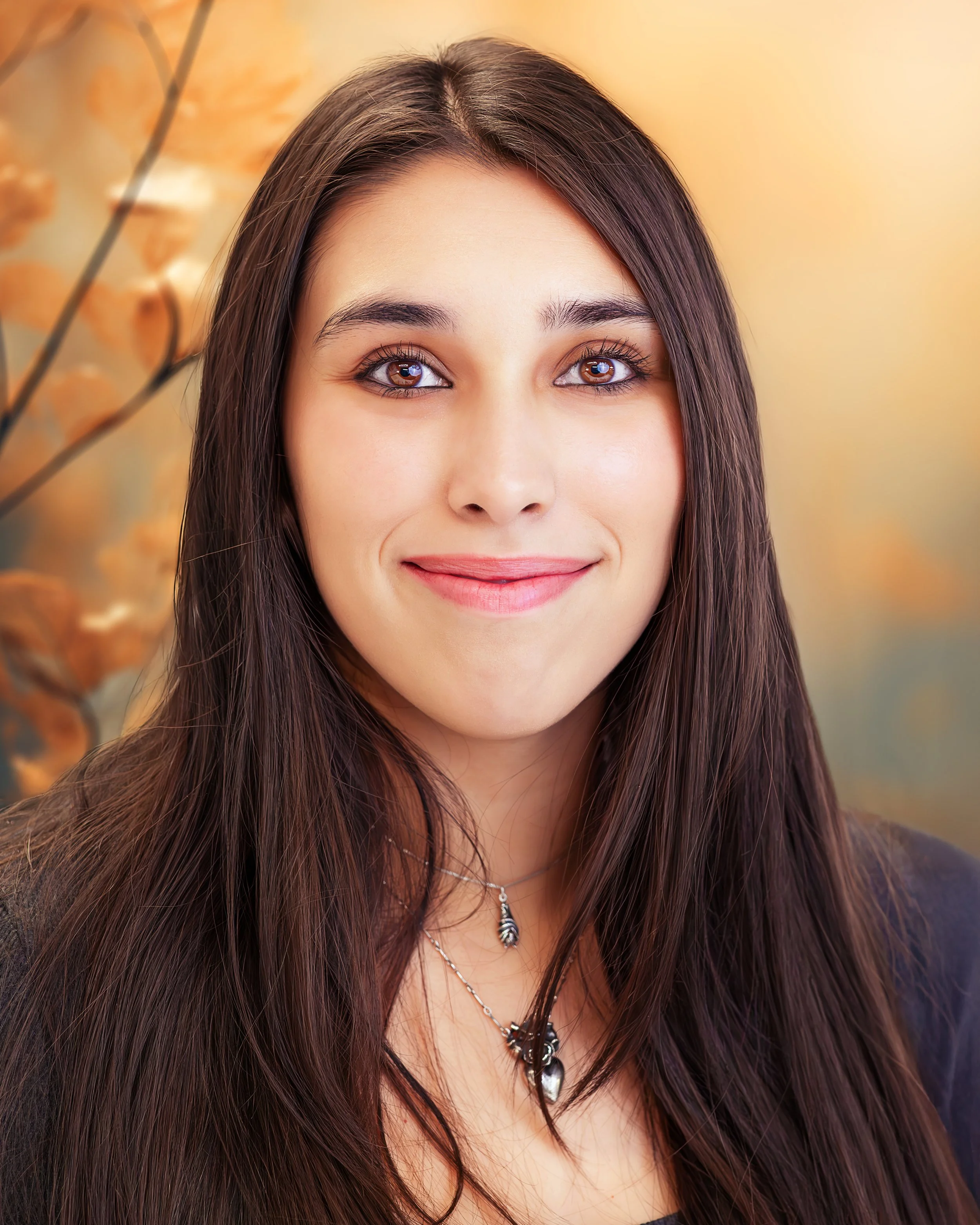 Close-up portrait of a young woman with long brown hair, brown eyes, and light skin, smiling gently, with a blurred autumnal background.