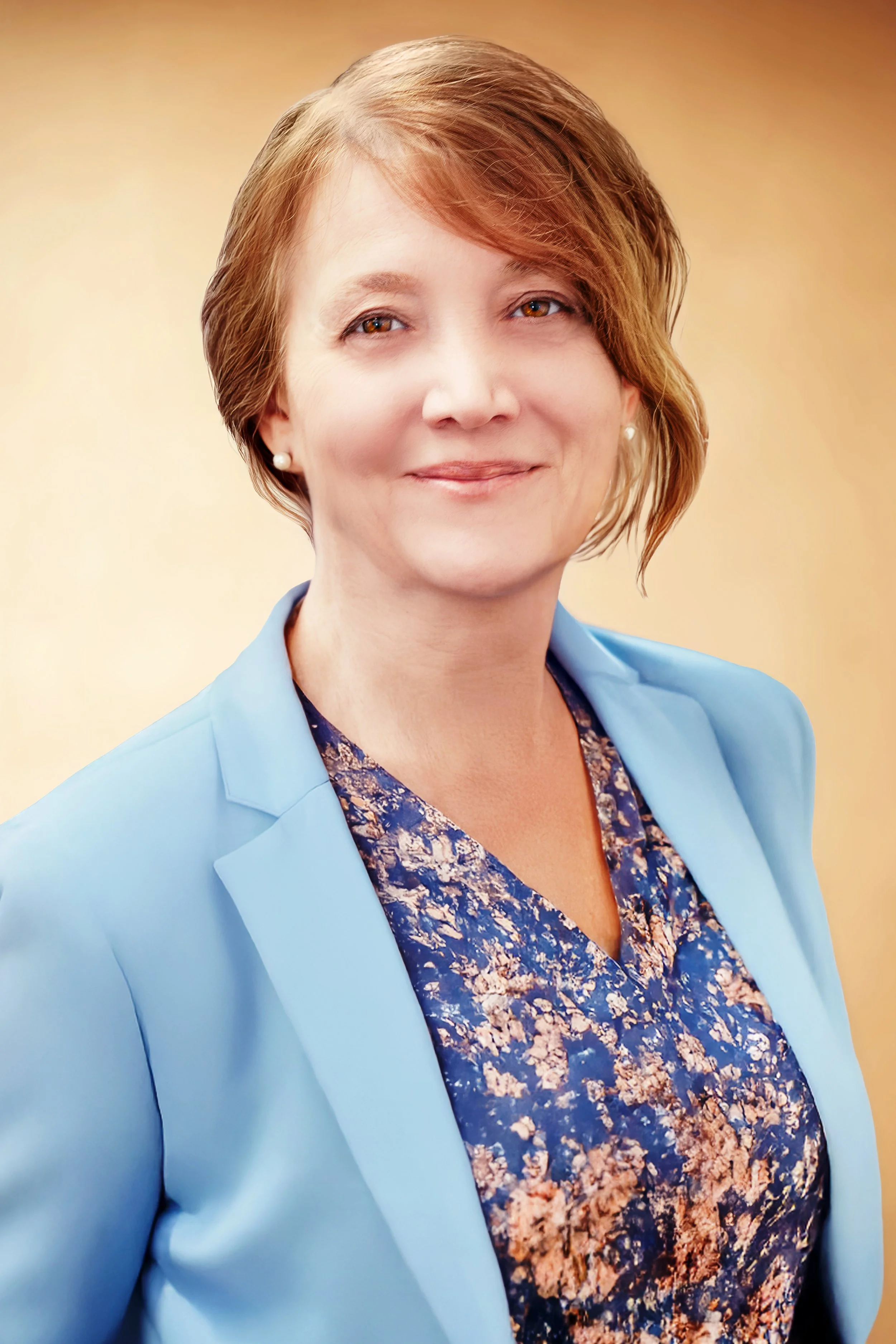 A woman with short light brown hair, wearing a light blue blazer over a patterned blouse with blue, pink, and beige colors, smiling against a beige gradient background. Headshot Photography in Loveland, Colorado. Jeanette Fellows Photography www.fell