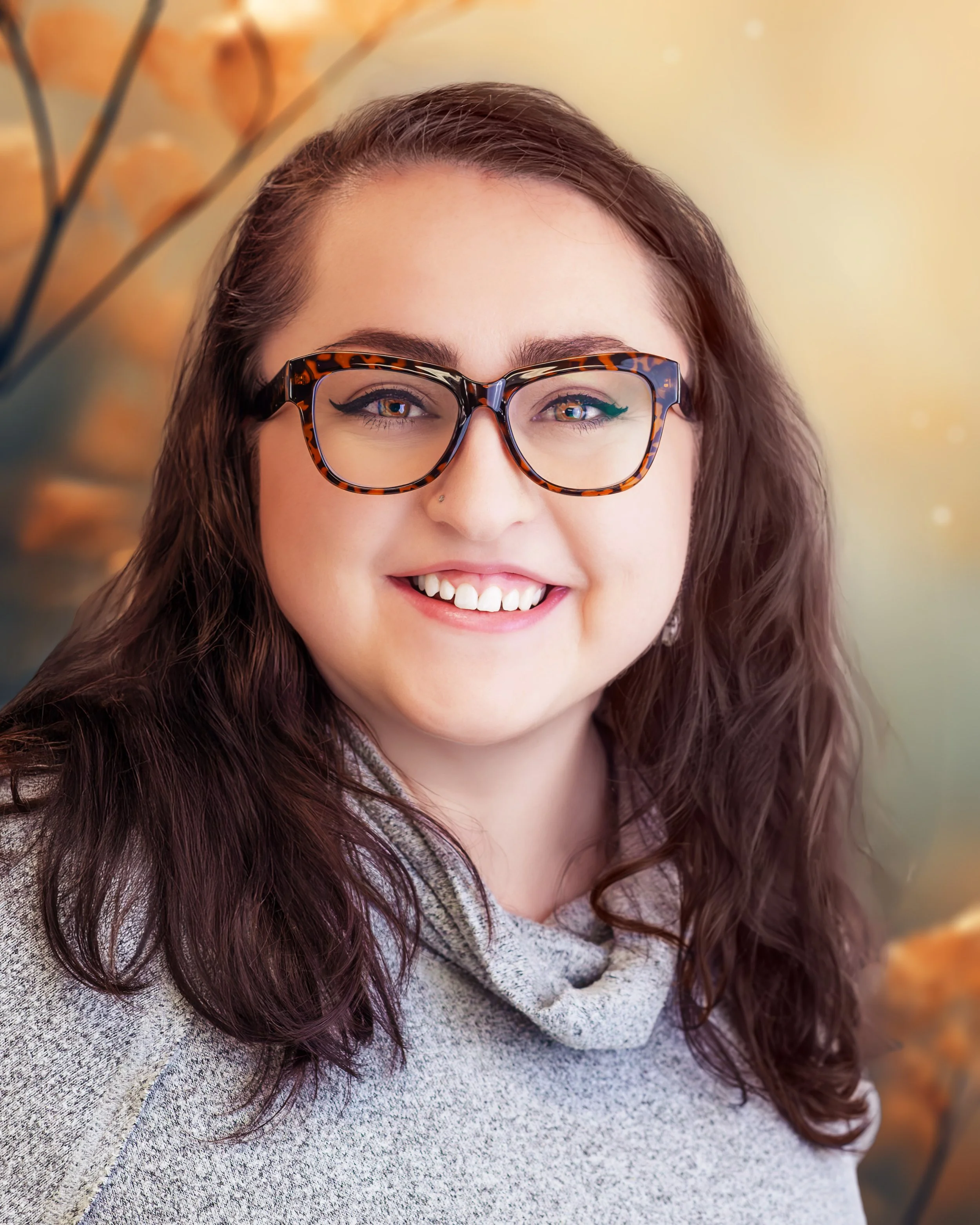 Portrait of a smiling woman with brown, curly hair, glasses with tortoiseshell frames, and a gray sweater, standing against a blurred background of orange and green fall foliage.