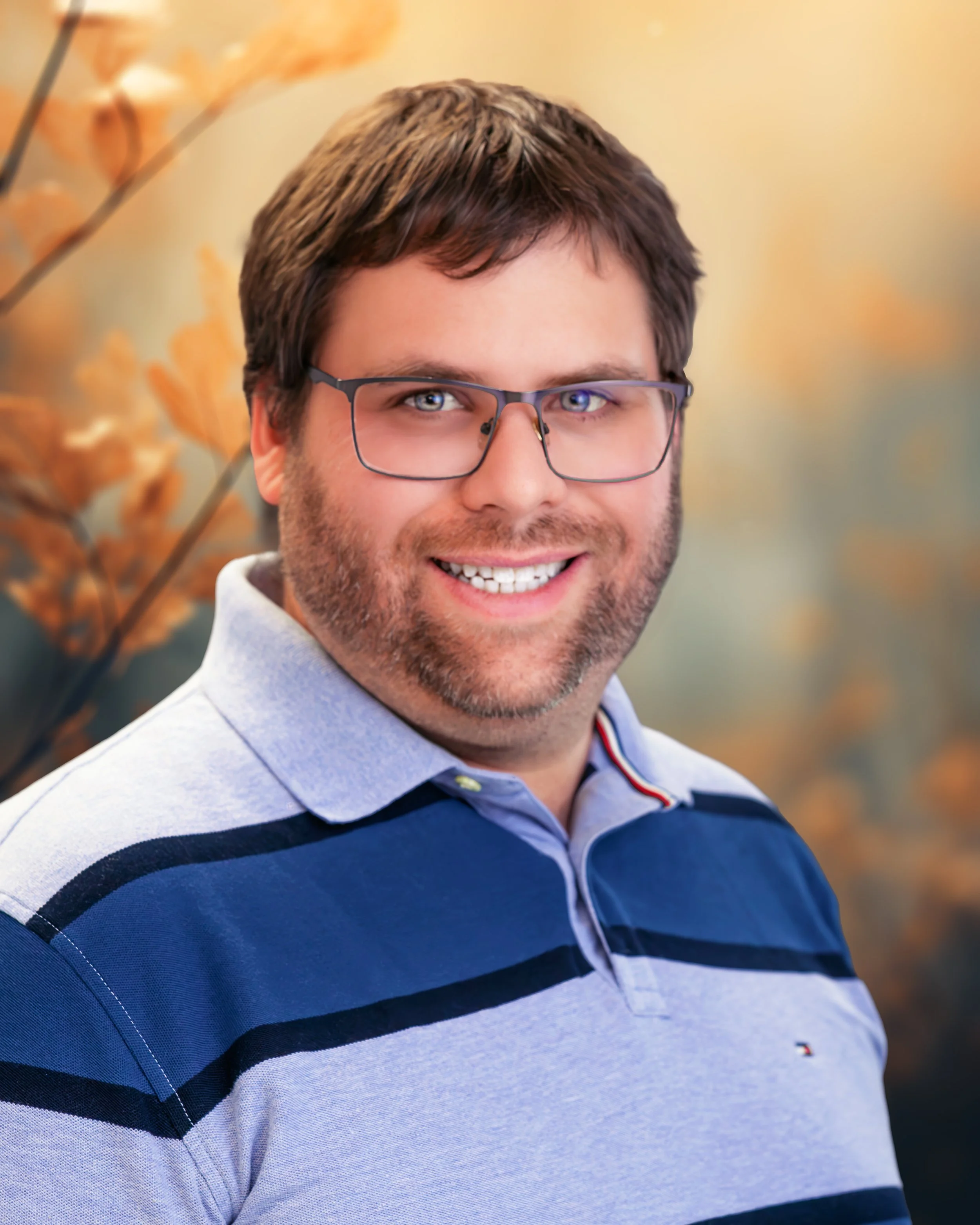 Portrait of a smiling man with glasses and brown hair, wearing a light blue polo shirt with navy and black stripes, against a blurred background of autumn leaves.