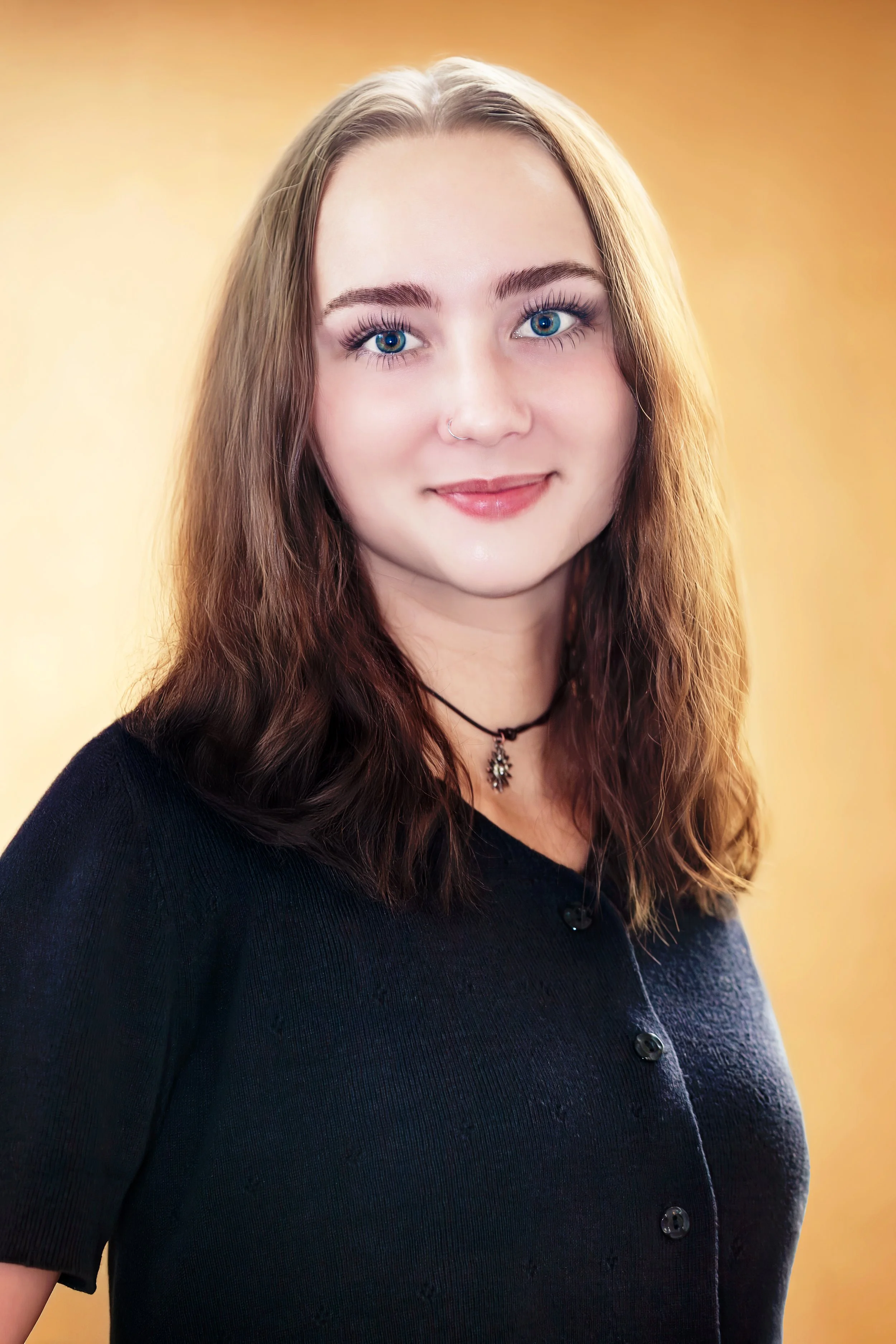 A young woman with blue eyes, light makeup, and a nose piercing, wearing a black button-up shirt and a choker necklace, smiling with a warm yellow background. Headshot Photography in Loveland, Colorado. Jeanette Fellows Photography www.fellows.photos