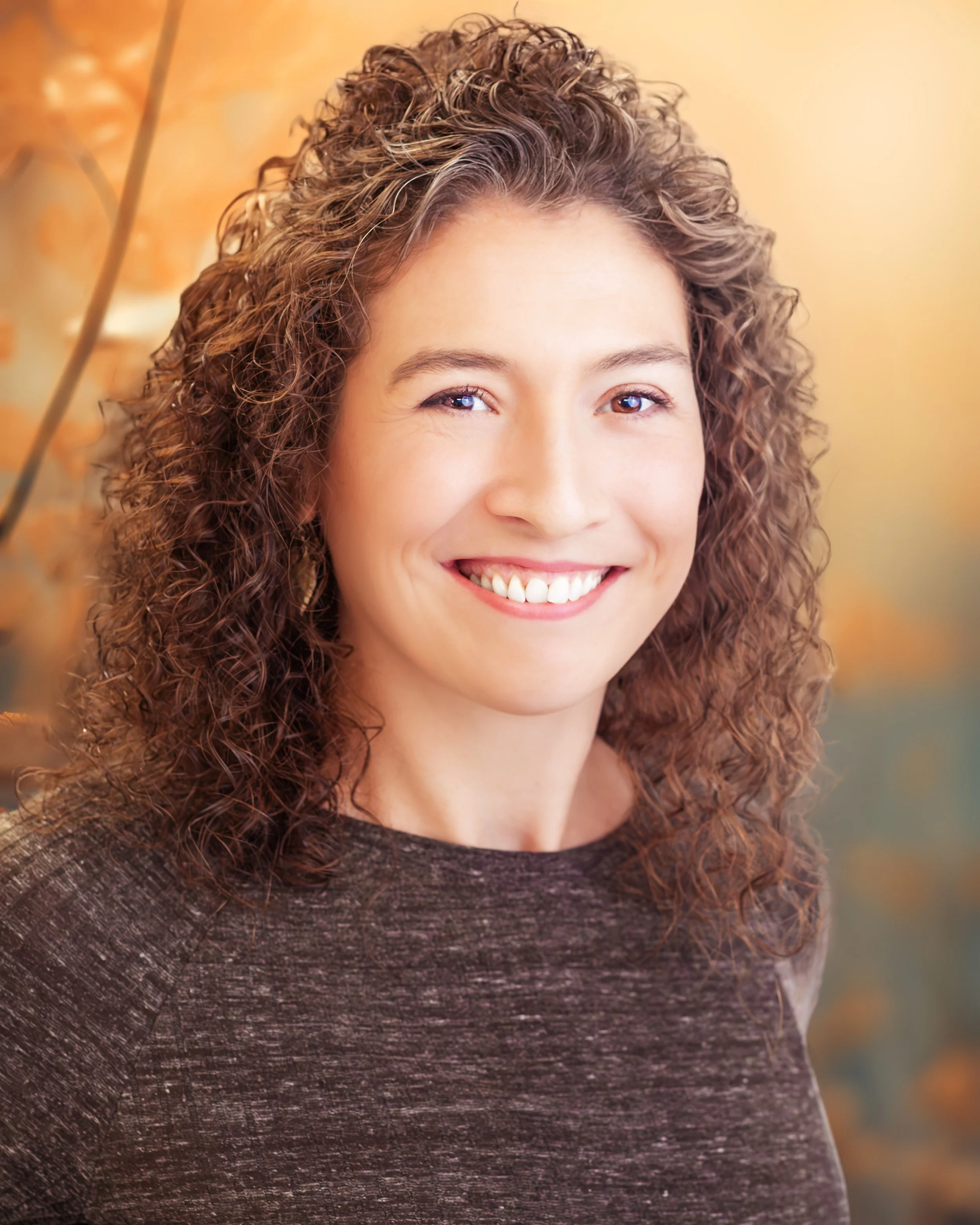 A woman with curly brown hair and a bright smile, wearing a dark textured top, standing outdoors with autumn-colored foliage in the background.