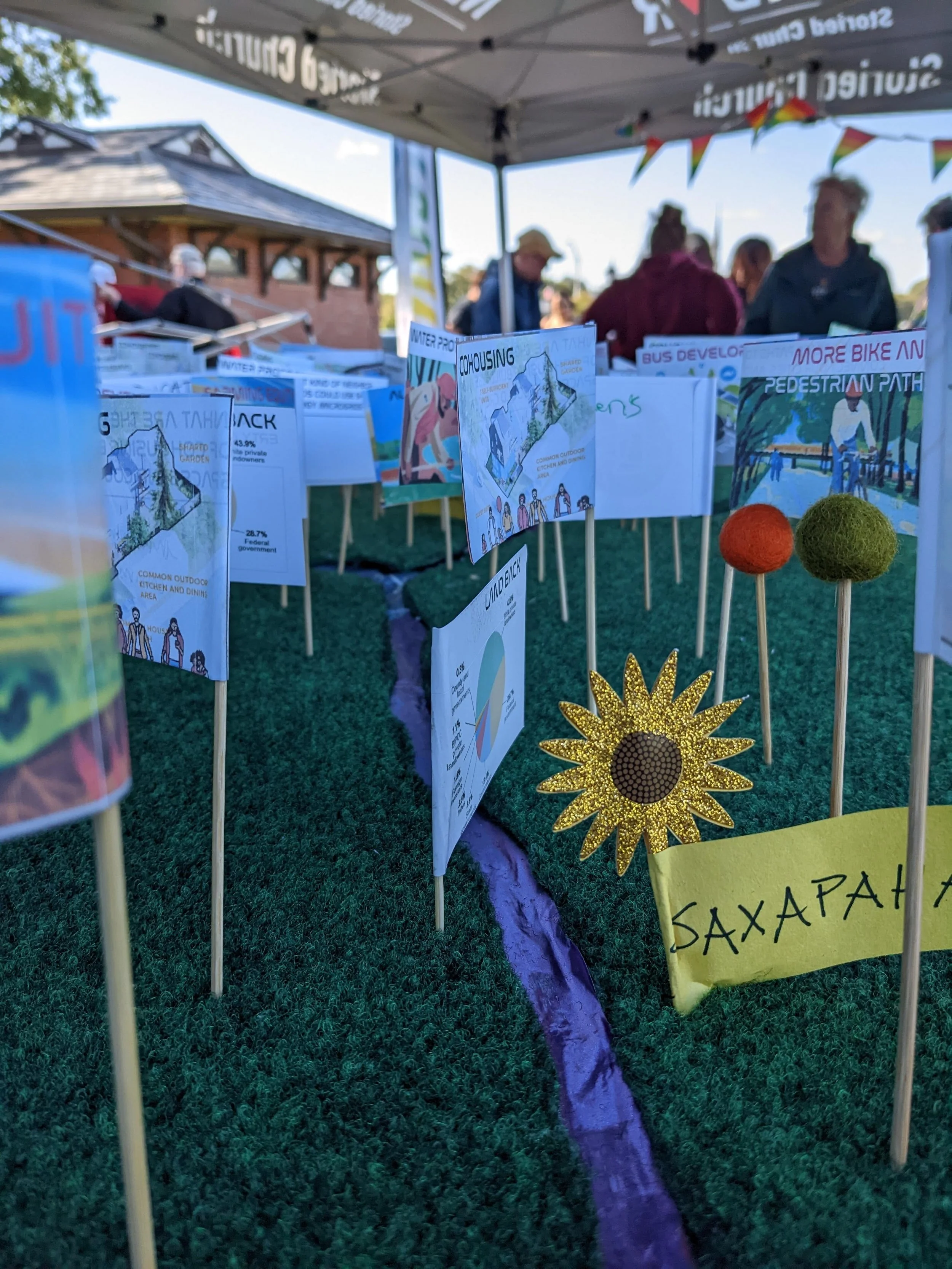Outdoor event exhibit with posters and artwork related to urban planning and community development. The display includes diagrams, graphs, and colorful decorations, with people gathering and talking under tents.
