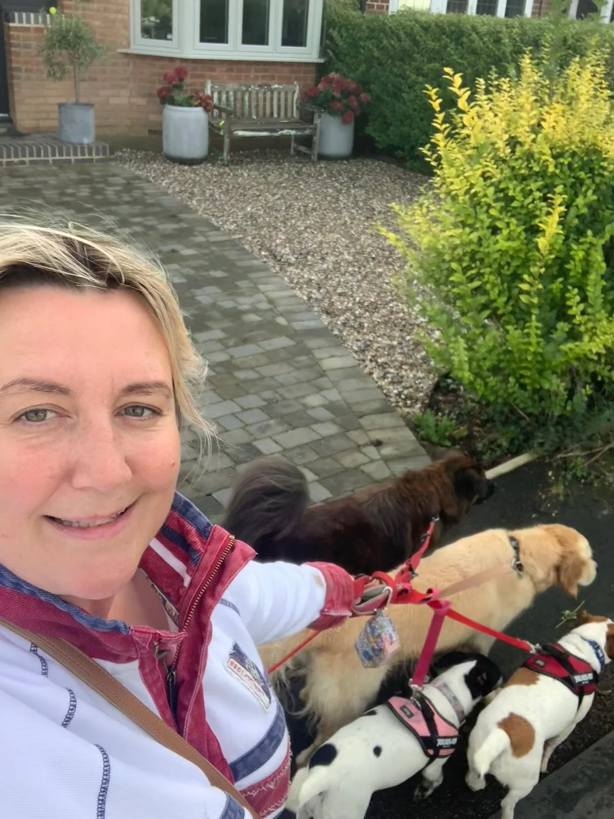 A woman takes a selfie with four dogs on leashes outside a house. The background shows a brick house, a garden bench, potted plants, and shrubs.