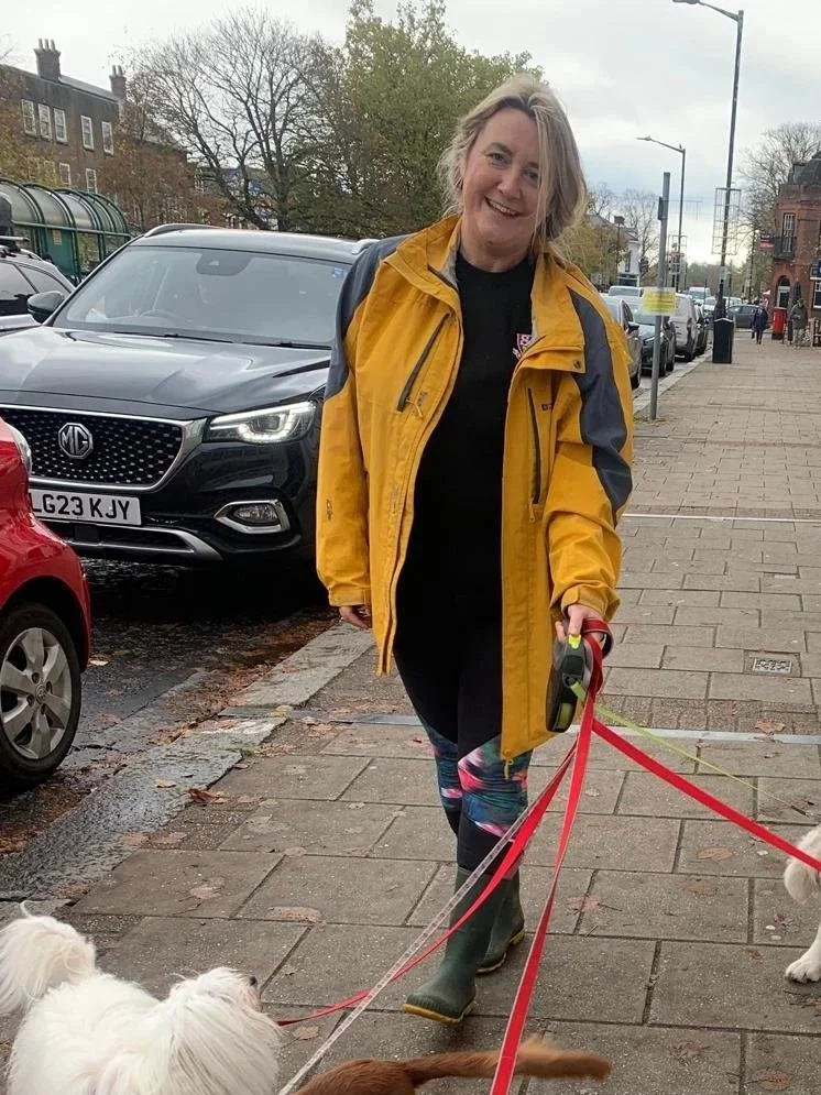 Woman walking dogs on a city sidewalk, wearing a yellow jacket, black outfit, colorful leggings, and rubber boots, smiling at camera with parked cars and buildings in the background.