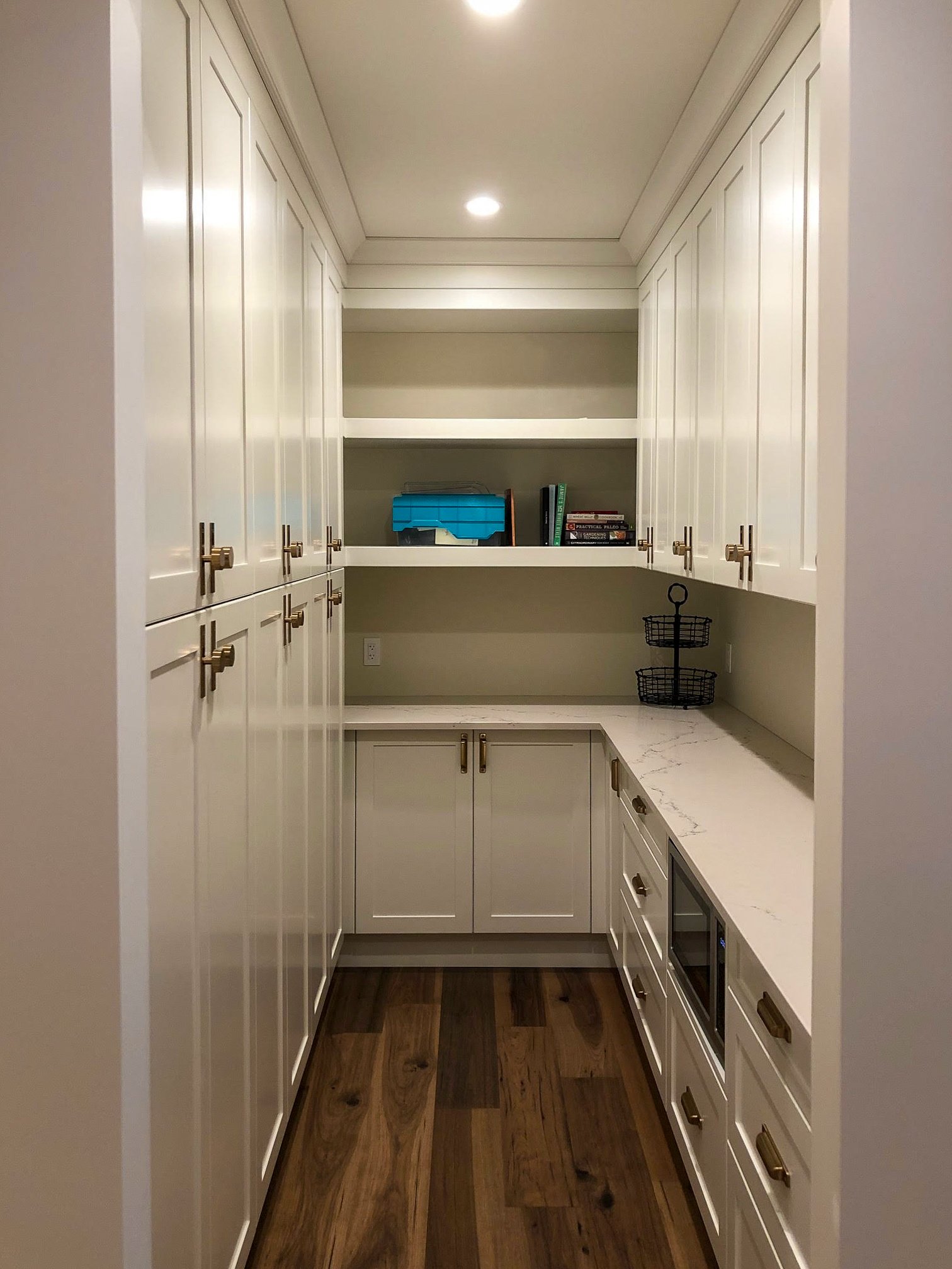 Modern pantry with white cabinets, shelves with books and containers, and hardwood flooring.
