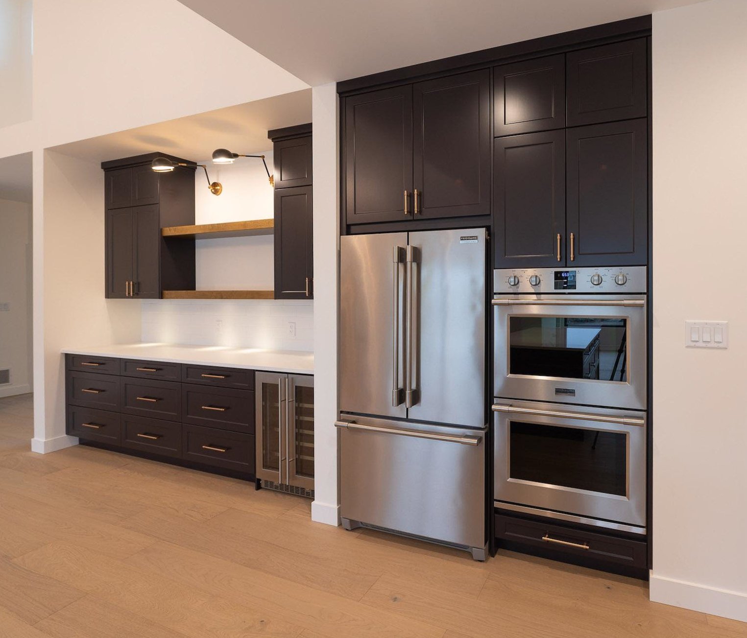 Modern kitchen with dark cabinets, stainless steel refrigerator and ovens, hardwood floor, and under-cabinet lighting.