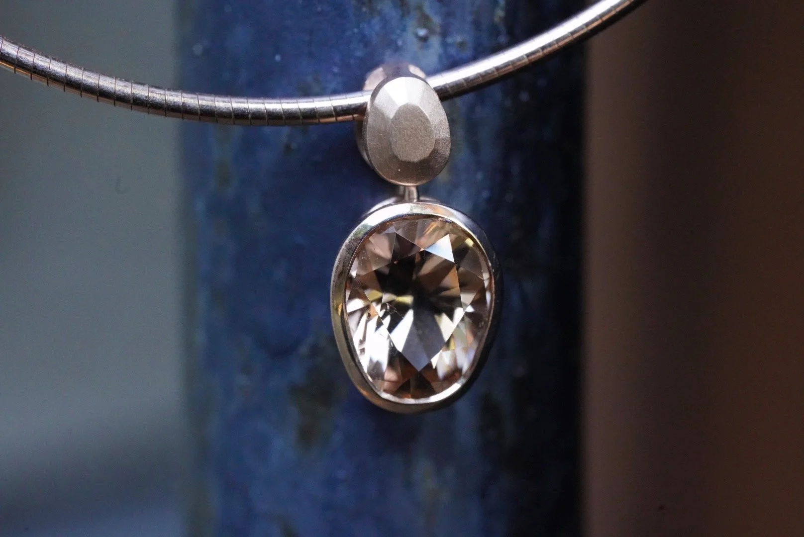 A rose gold necklace with an egg shaped, honey-golden tourmaline strung on an omega chain against a steely black background
