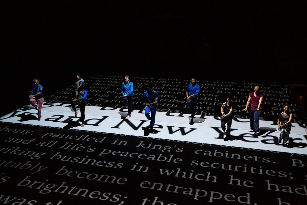 Nine dancers in athletic clothing perform on a stage with a floor covered in large white and black text from a newspaper or book.