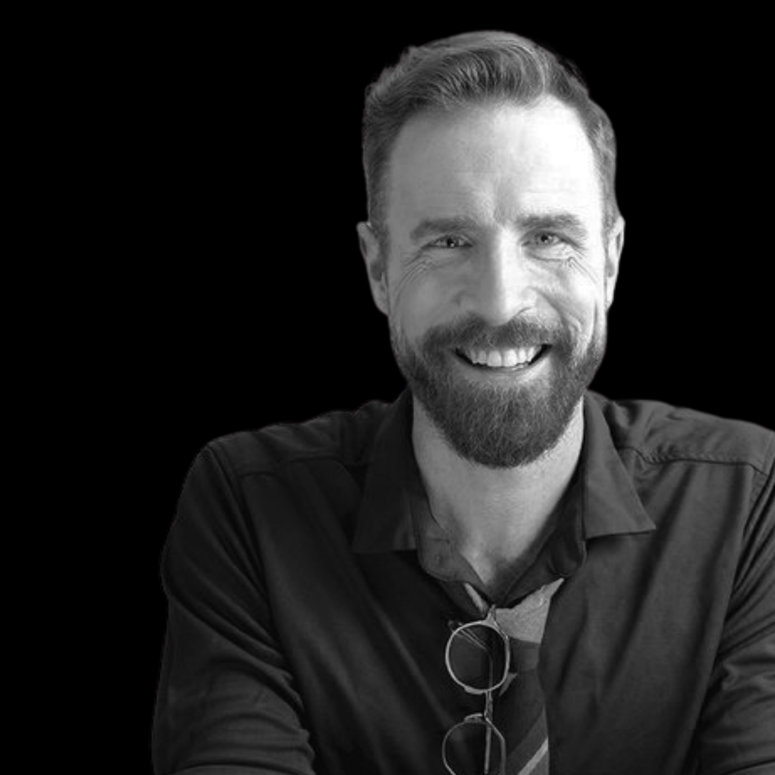 Black and white photo of a smiling man with a beard and short hair, wearing a collared shirt and sunglasses hanging from his shirt.