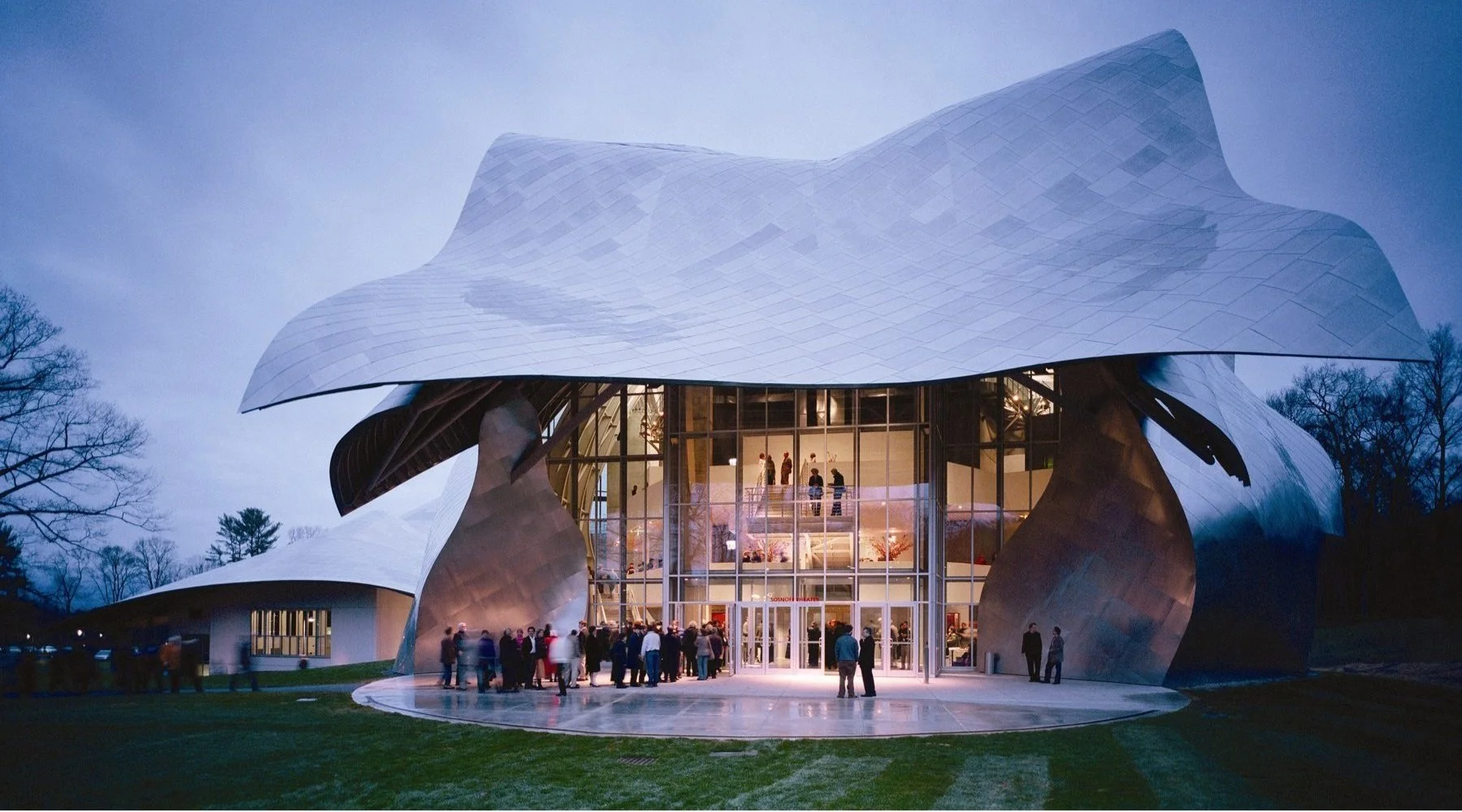 Modern cultural or concert hall with an organic, flowing metallic roof and large glass entrance, surrounded by trees, with people gathering outside in the evening.