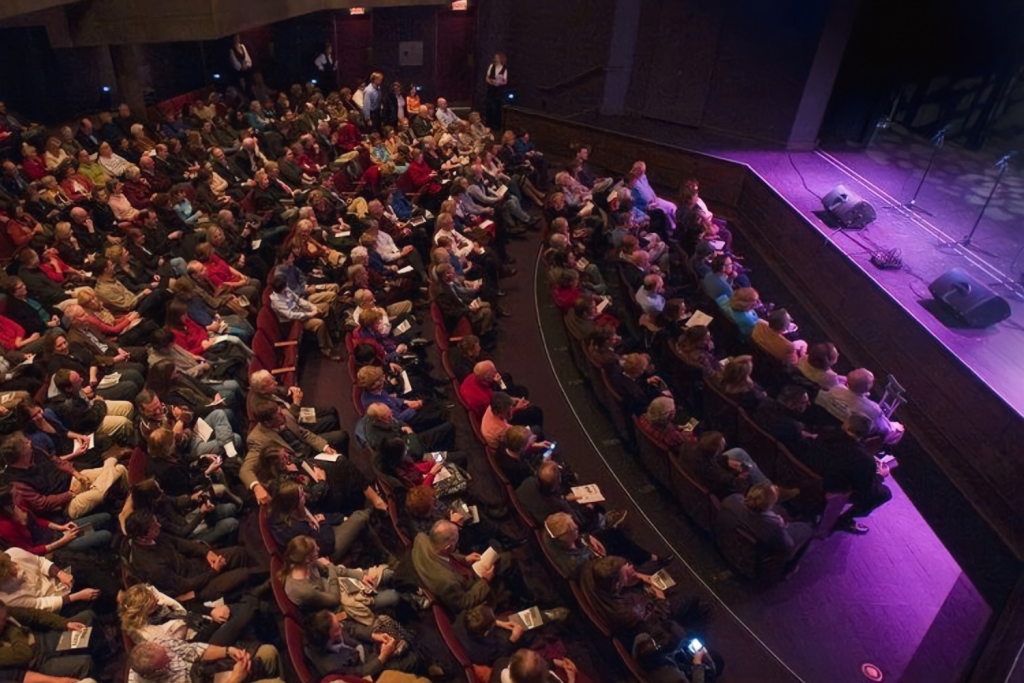 Large audience seated in a theater, waiting for a performance or event to start, with purple stage lighting and minimal stage setup.