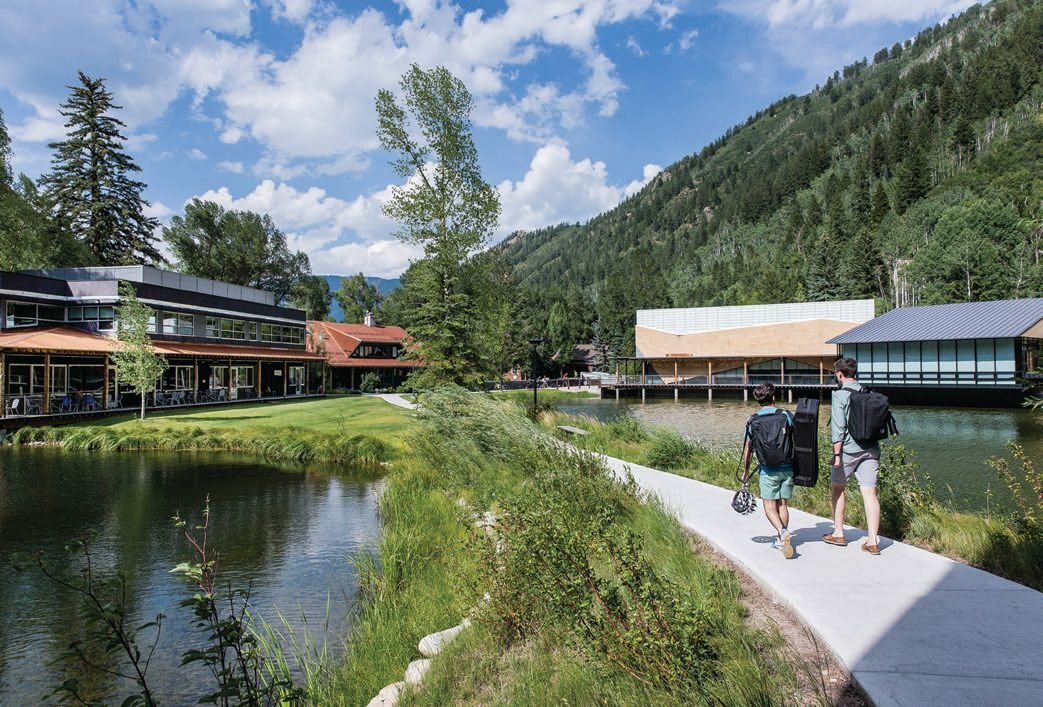 Two young men with backpacks walk on a paved path beside a river, surrounded by lush greenery and mountains, with modern buildings in the background on a bright, partly cloudy day.