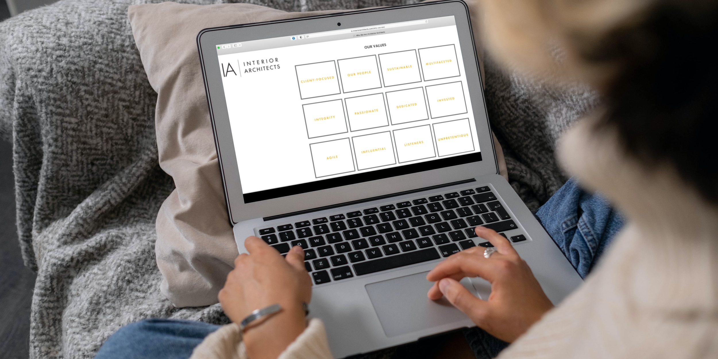 A person sitting on a couch using a laptop, which displays a webpage with values of an interior architecture firm.