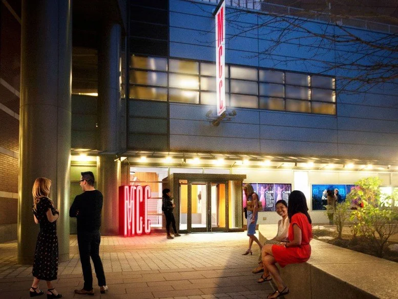 People gathered outside a building with an illuminated sign that reads 'MGC' and a vertical sign above the entrance, at night.