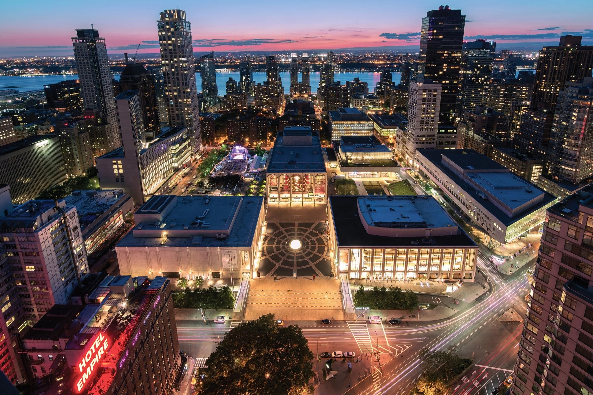 Aerial view of a city skyline at dusk with tall buildings, city streets, and a river in the background. The streets are illuminated with traffic lights and moving cars, and the sky displays pink and purple hues.