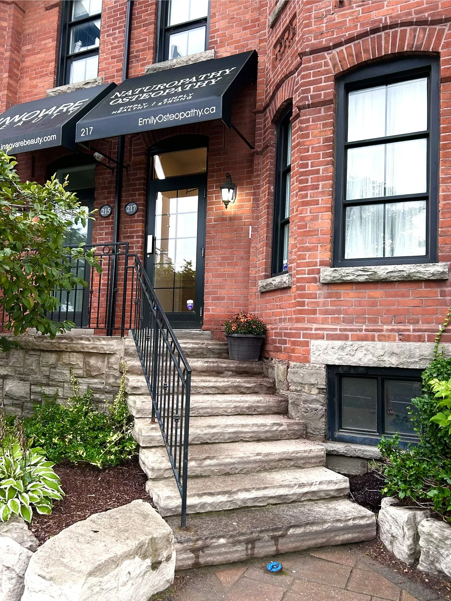 A brick building with a black door and large windows. There are stone stairs leading up to the door, with a black metal railing on the stairs. To the right of the stairs, there is a large flowerpot with flowers. The building has a black awning with white text that reads 'NATUROPATHY & OSTEOPATHY' and 'Emily Osteopathy.ca' and the number 217.