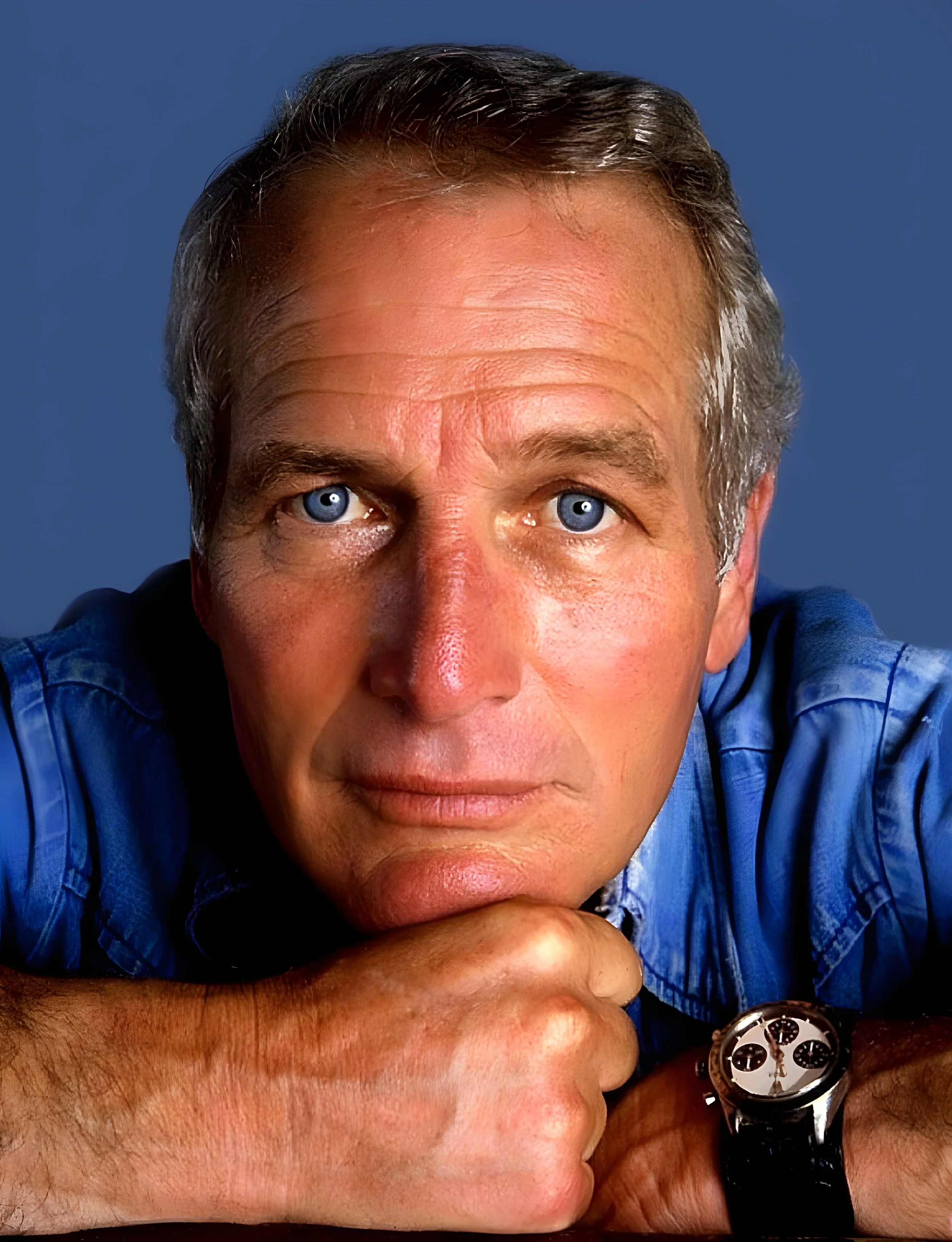 Close-up of a middle-aged man with blue eyes and gray hair, resting his chin on his arm, wearing a blue shirt and a wristwatch, against a blue background.