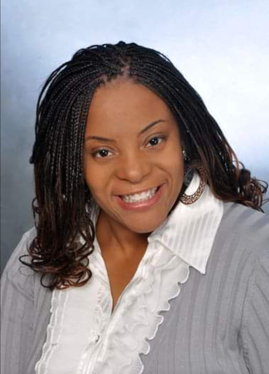 A woman with shoulder-length curly hair styled in braids, smiling and wearing a white shirt with ruffled edges and earrings, against a neutral background.