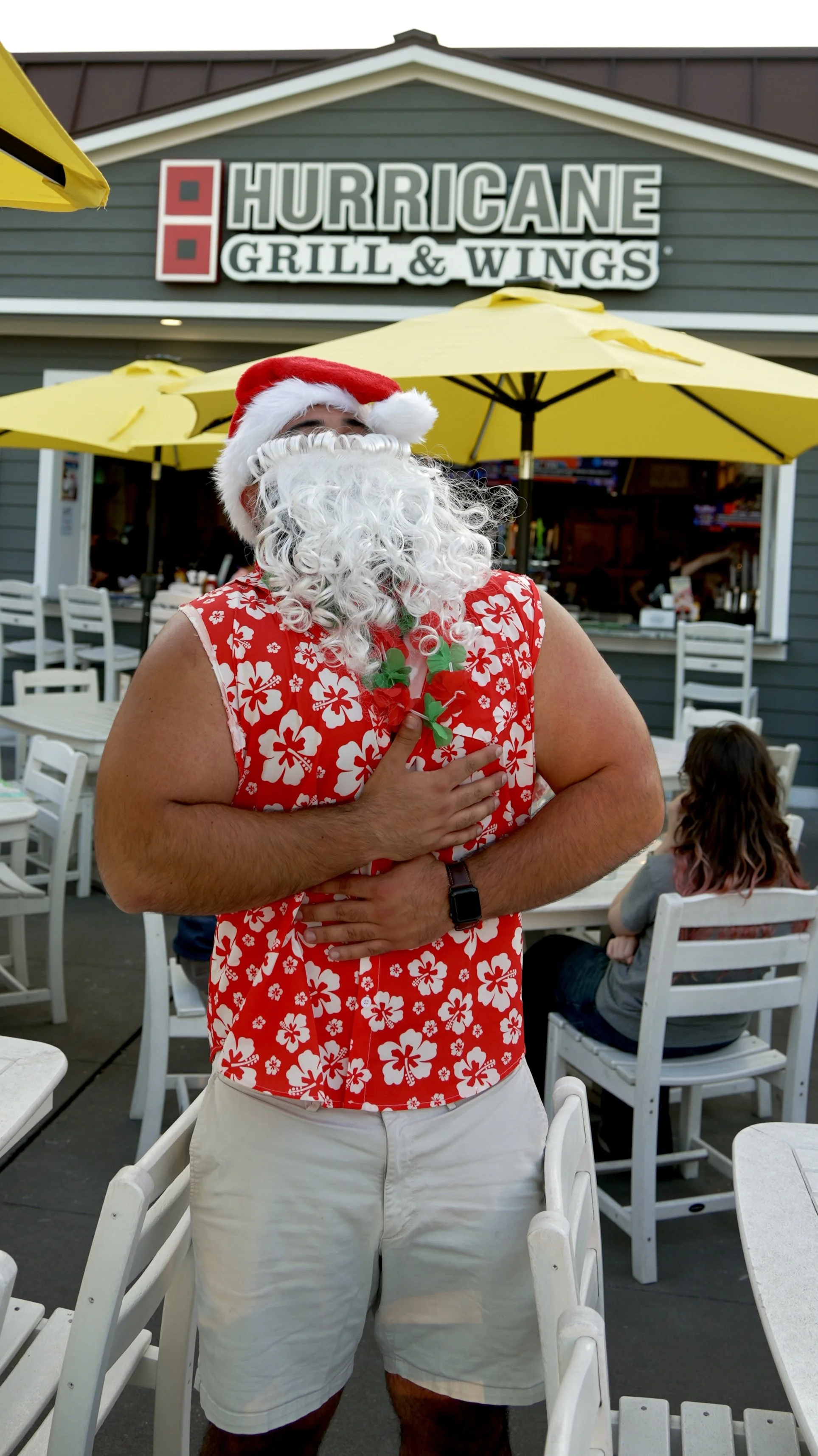 A man wearing a Santa hat, curly white beard, and a red Hawaiian shirt with white flowers, standing outside at a restaurant called Hurricane Grill & Wings. He is holding his hand over his chest and appears to be singing or speaking.
