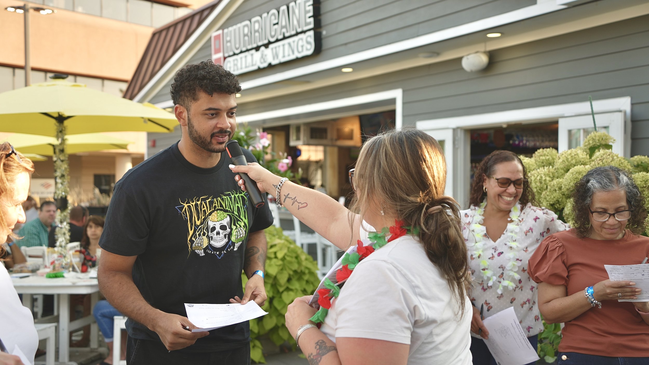 People gathered outdoors in front of a restaurant called Hurricane Grill & Wings. A man with curly hair and a beard is holding a microphone and a paper while talking to a woman wearing white with a flower lei. Several women are smiling and holding pa
