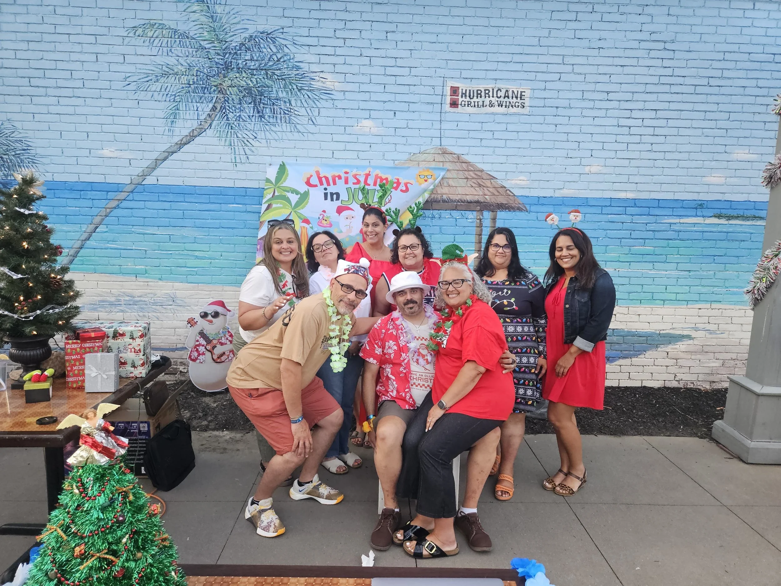 A group of eight people gathered outdoors in front of a mural with a palm tree, cloud, and beach scene, celebrating Christmas in July, with Christmas decorations and gifts around them.