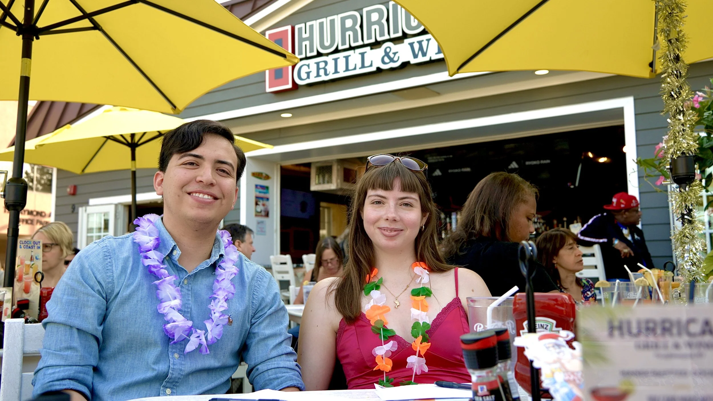 Two people smiling and sitting at an outdoor restaurant under yellow umbrellas, decorated with leis, with a Taco Burrito Grill sign visible above.