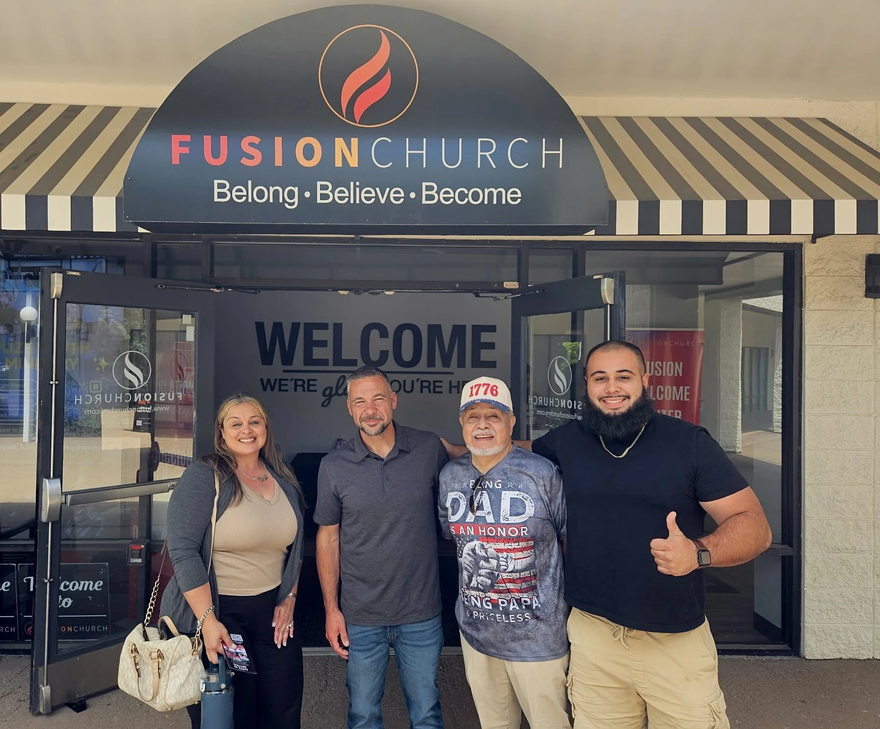 A group of five people standing in front of Fusion Church entrance, smiling, with a woman on the left holding a beige handbag, next to a man in a dark gray shirt, an older man wearing a cap in the middle, a young man with a beard on the right giving 