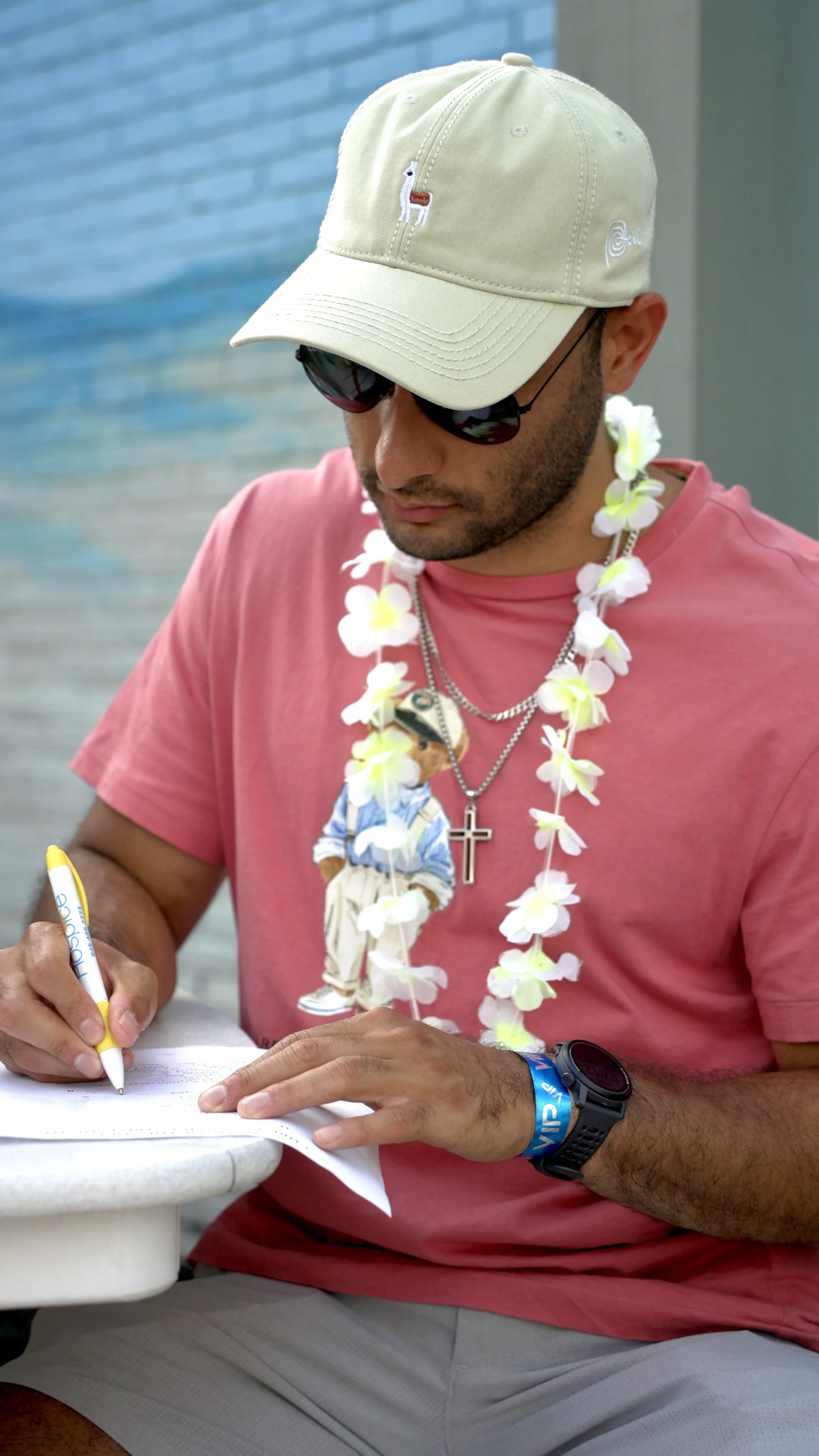 A man wearing a cream baseball cap, sunglasses, a pink t-shirt, and a lei with flowers is signing a paper with a yellow and white marker. He is wearing a watch and a blue wristband.