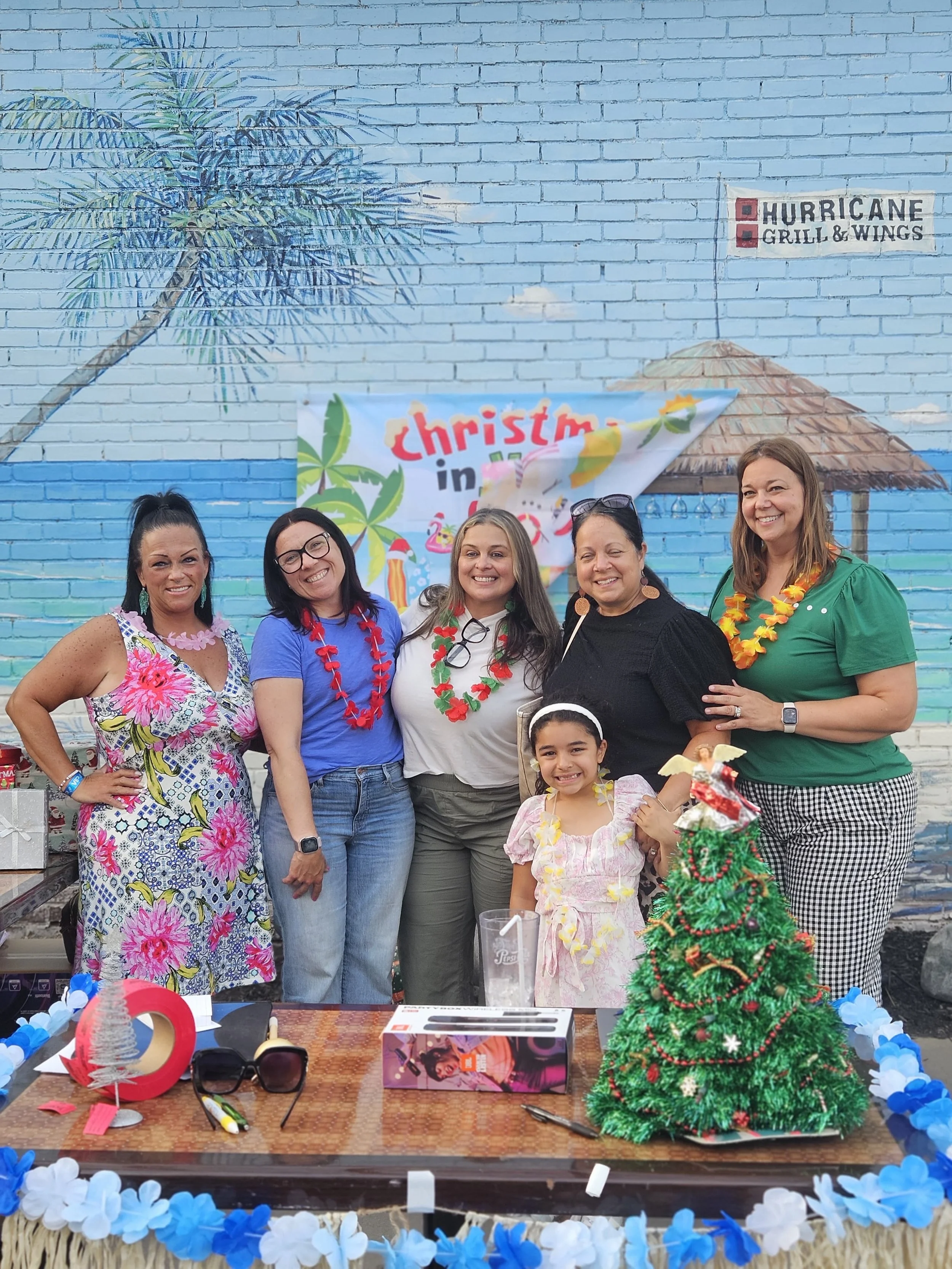 Group of five women and one young girl smiling at a festive outdoor party with tropical and holiday decorations. A blue brick wall with a painted mural of a palm tree and a hut is in the background, and a sign for 'Hurricane Grill & Wings' is visible