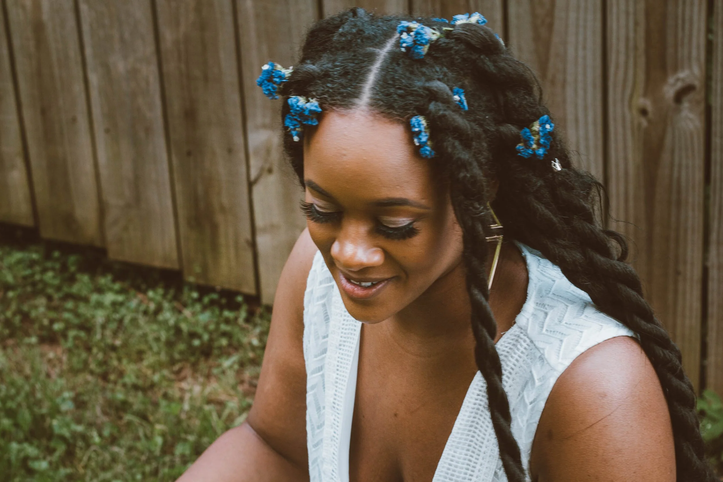 A woman with dark, textured hair styled in dreadlocks decorated with small blue flowers, smiling gently, wearing a white sleeveless top, with a wooden fence and grass in the background.