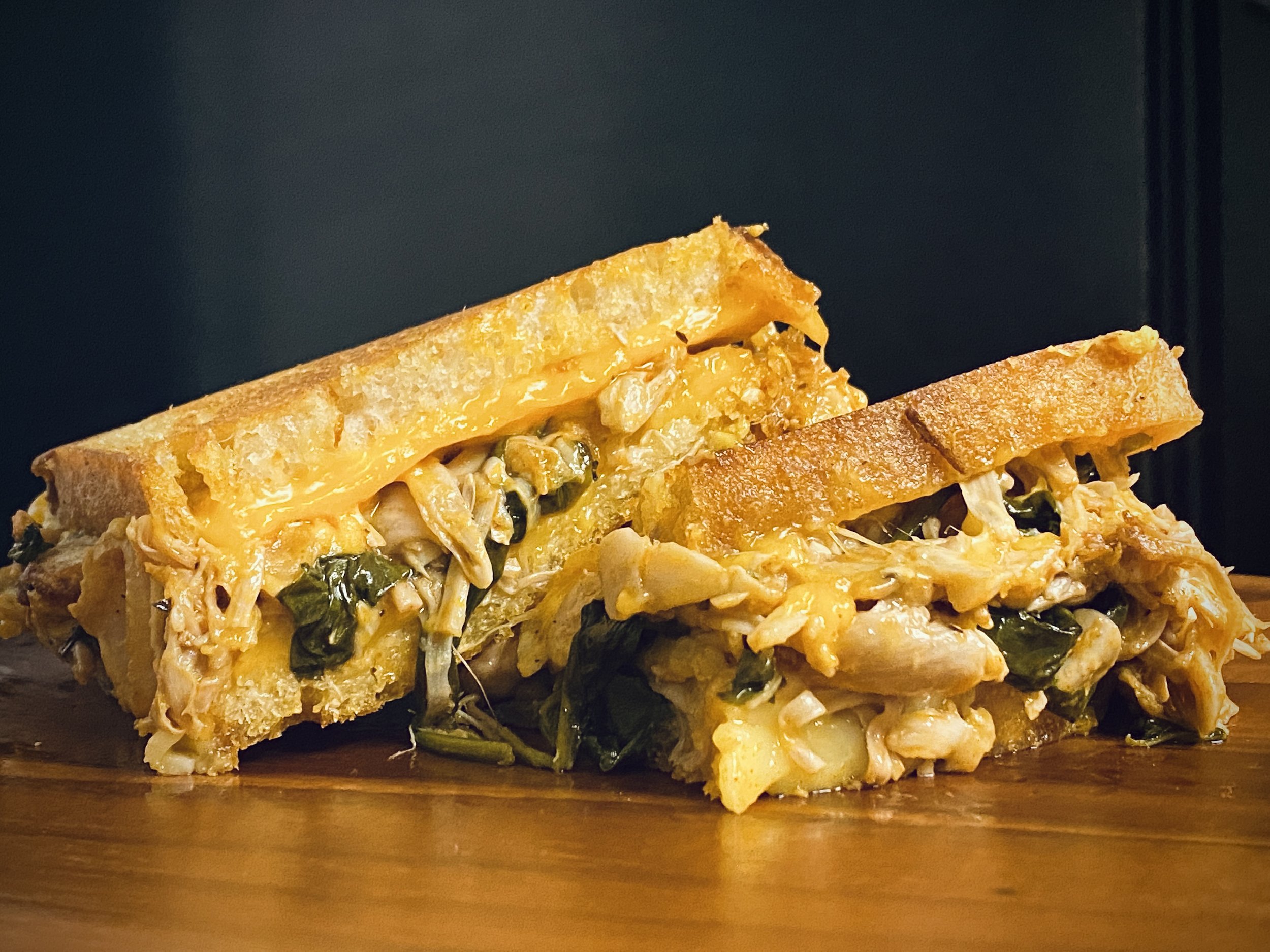 Close-up of a grilled cheese sandwich with chicken, spinach, and melted cheddar cheese on a wooden surface.