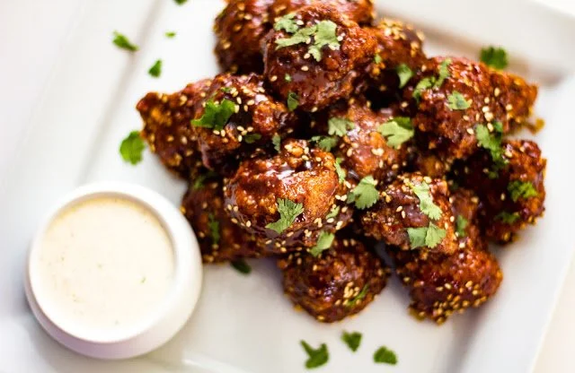 Fried cauliflower wings garnished with cilantro and sesame seeds, served with a side of dipping sauce on a white plate.