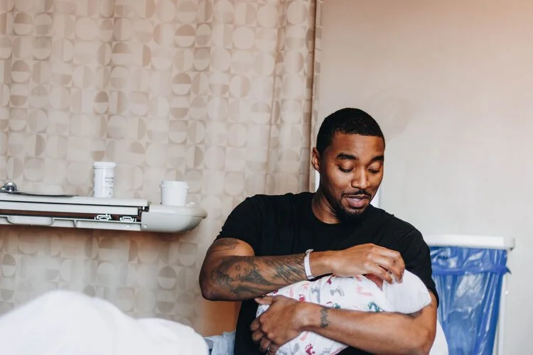 A man holding a newborn baby in a hospital room with medical supplies and a blue laundry basket in the background.